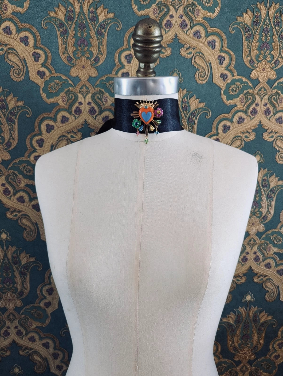 Broochella Sacro cuore apolloinia multicoloured enamel heart shaped brooch. Pinned to a mannequin. The background is green and gold damask wallpaper