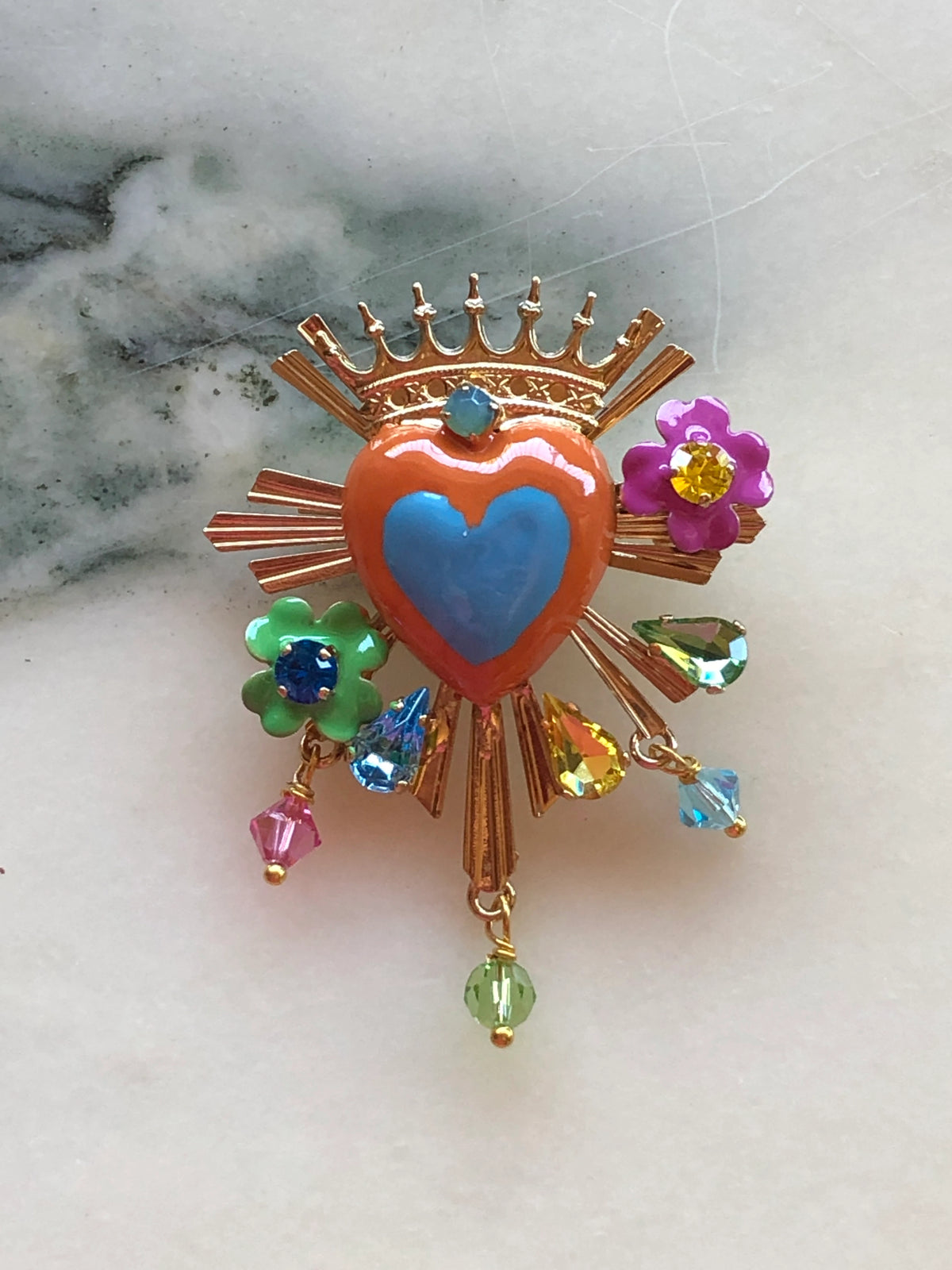 Broochella Sacro Cuore Apollonia Heart Brooch featuring a coral and blue enamel sacred heart, gold-tone crown and rays, colourful floral details, crystals and dangling beads, photographed on a white marble background.