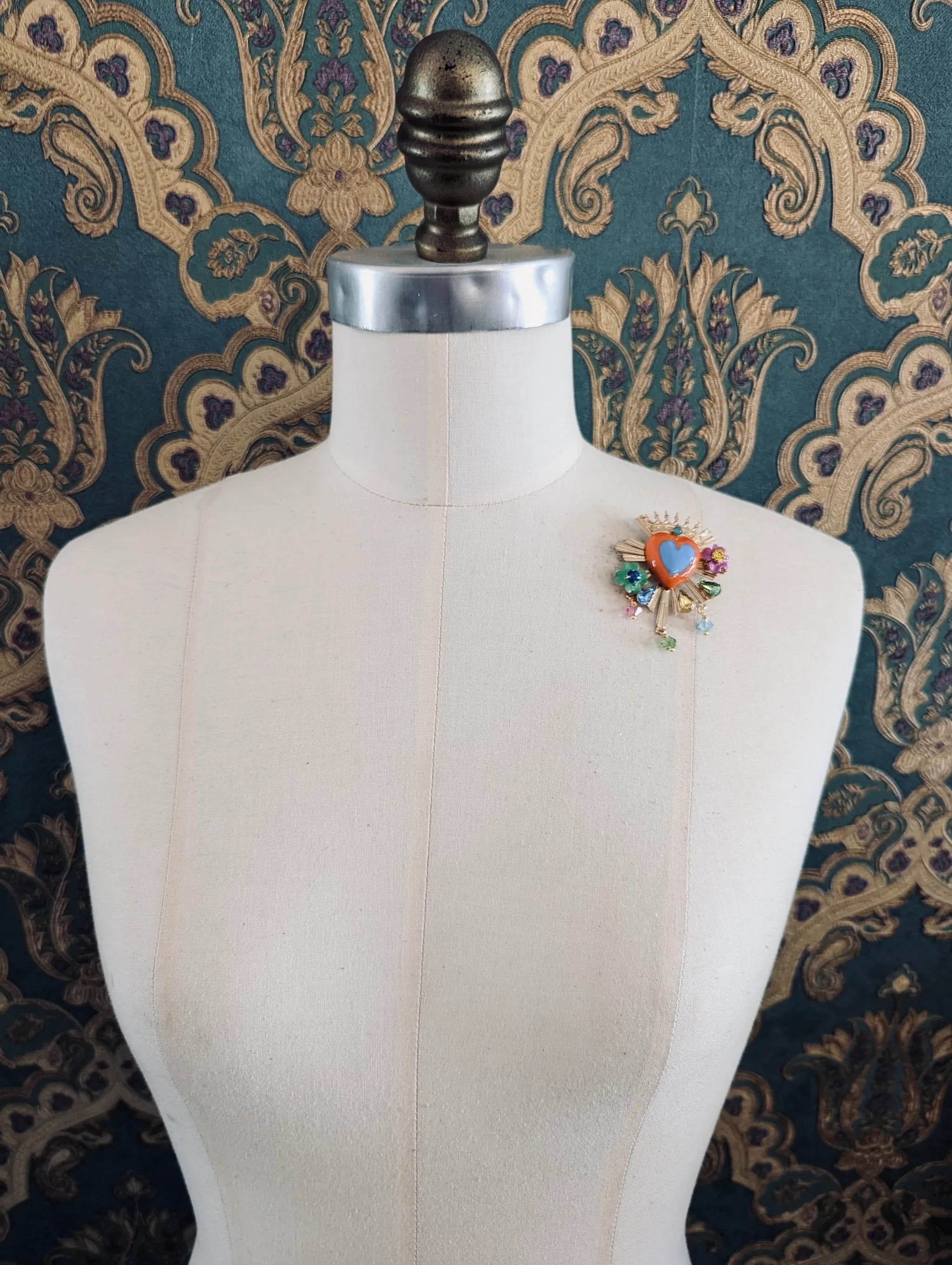Broochella Sacro cuore apolloinia multicoloured enamel heart shaped brooch. Pinned to a mannequin. The background is green and gold damask wallpaper