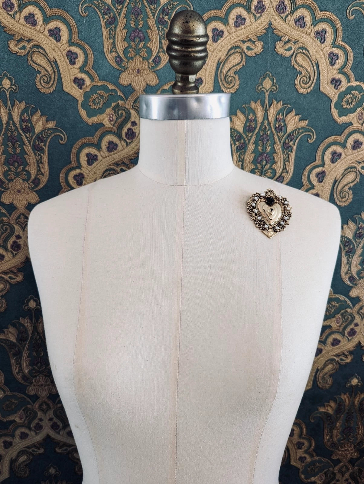 Broochella Sacro cuore benedetta black and gold metal  heart shaped brooch. Pinned to a mannequin. The background is green and gold damask wallpaper