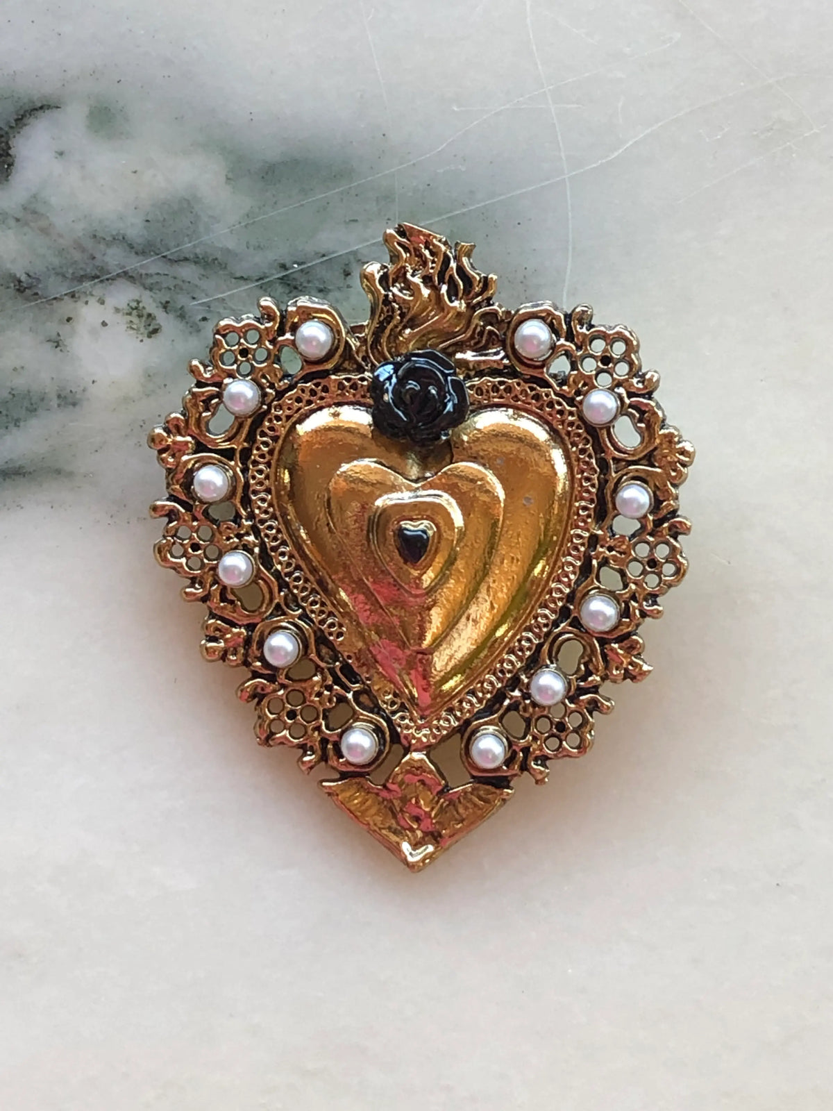 Broochella Sacro Cuore Benedetta Heart Brooch in red, featuring an ornate gold-tone sacred heart with red rose detail, pearl embellishments and flame motif, photographed on a white marble background