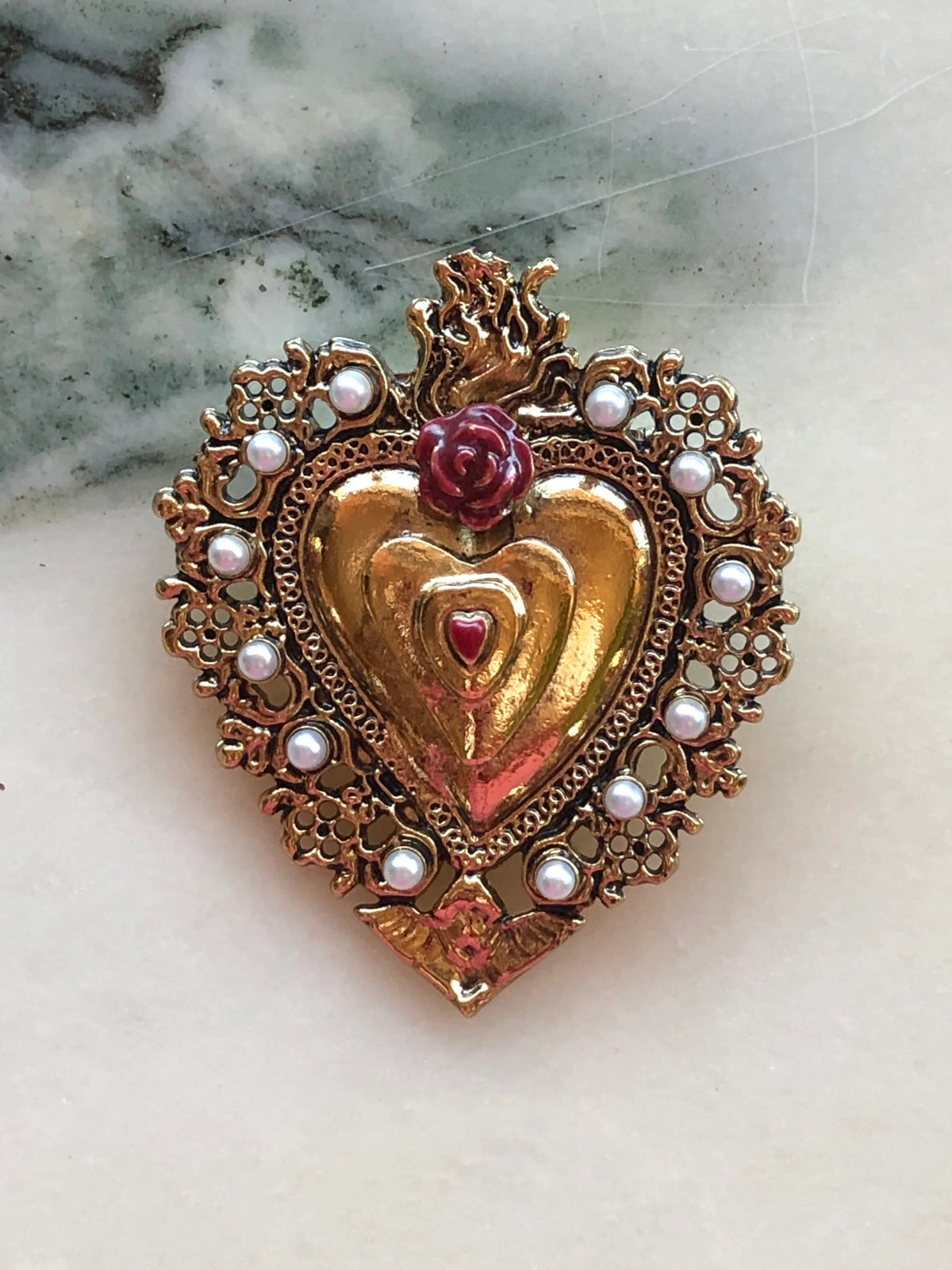 Broochella Sacro Cuore Benedetta Heart Brooch in red, featuring an ornate gold-tone sacred heart with red rose detail, pearl embellishments and flame motif, photographed on a white marble background