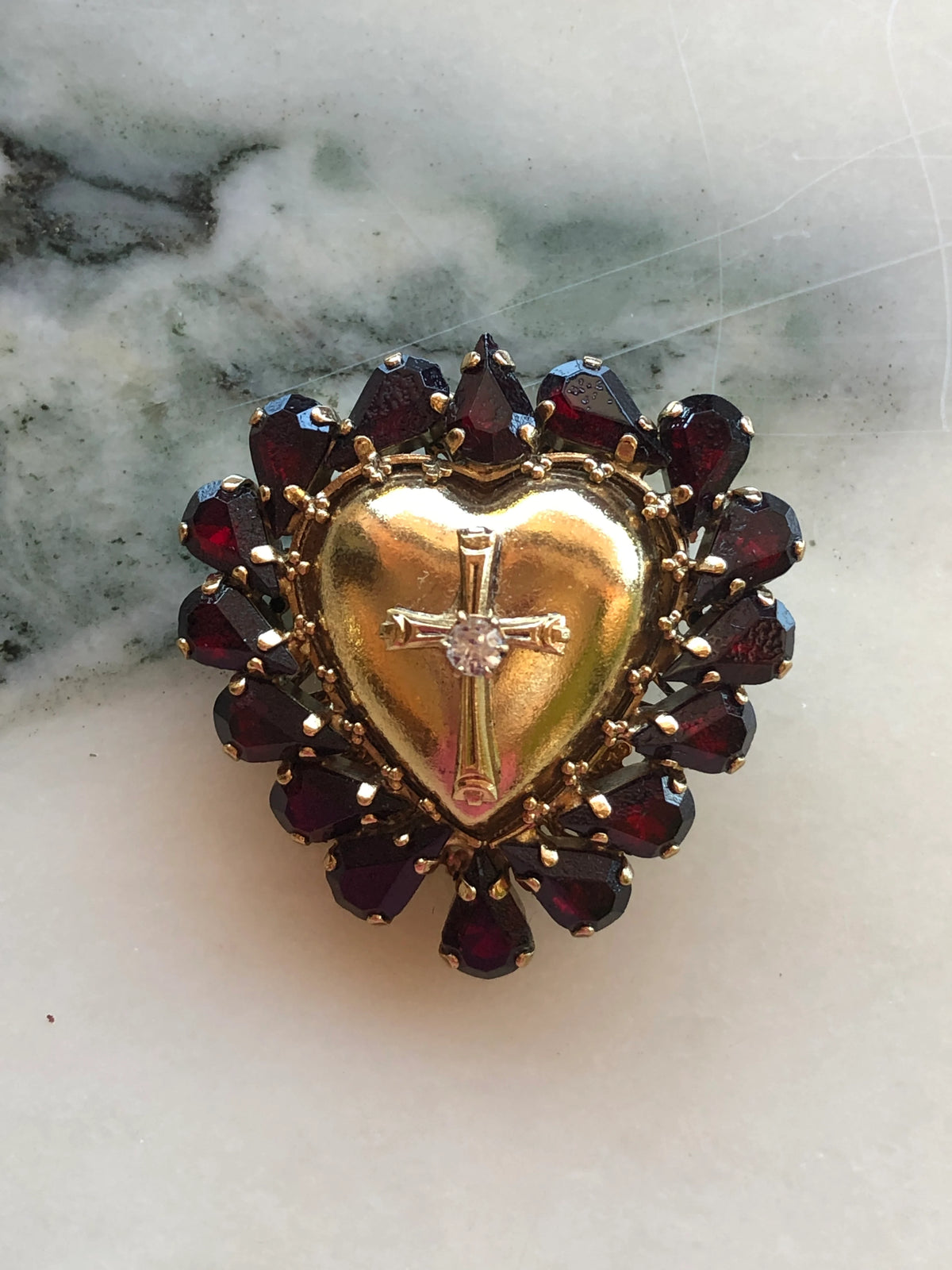 Broochella Sacro Cuore Cristo Heart Brooch featuring a gold-tone sacred heart with cross and crystal centre, surrounded by deep garnet red faceted stones, photographed on a white marble background.
