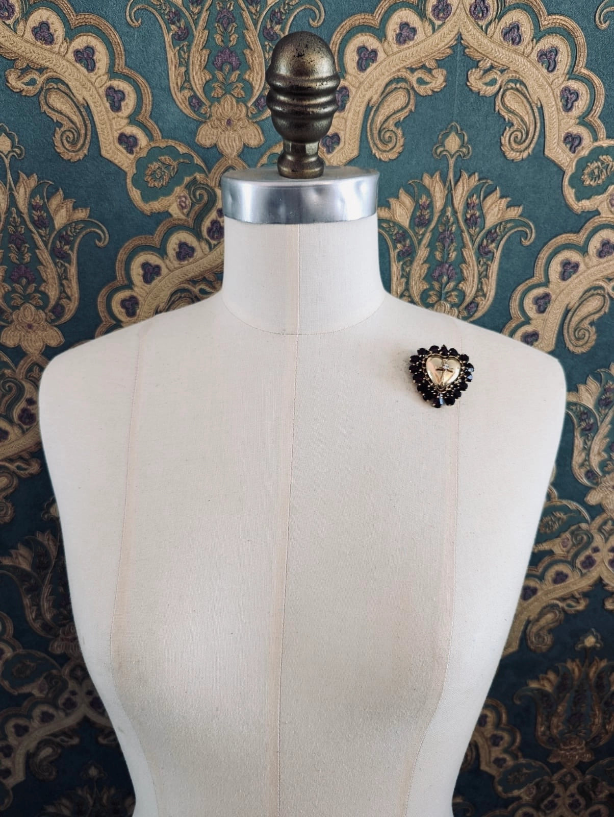 Broochella Sacro cuore cristo gold metal  heart shaped brooch, with a crucifix centre and a red crystal frame. Pinned to a mannequin. The background is green and gold damask wallpaper
