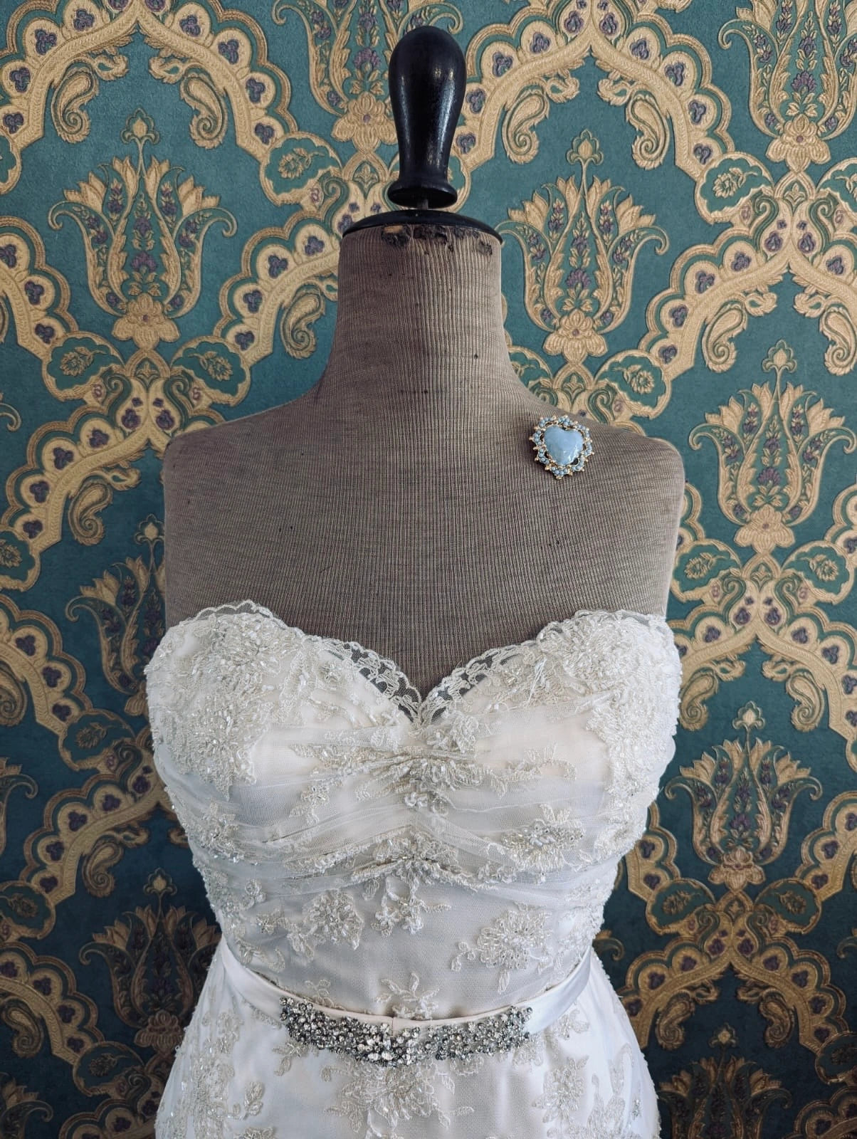 Broochella sacro cuore fiorentina blue bridal heart shaped enamel brooch with gold metal base. Pictured on a mannequin that is wearing a white, beaded strapless wedding dress. The background is green and gold damask wallpaper. 