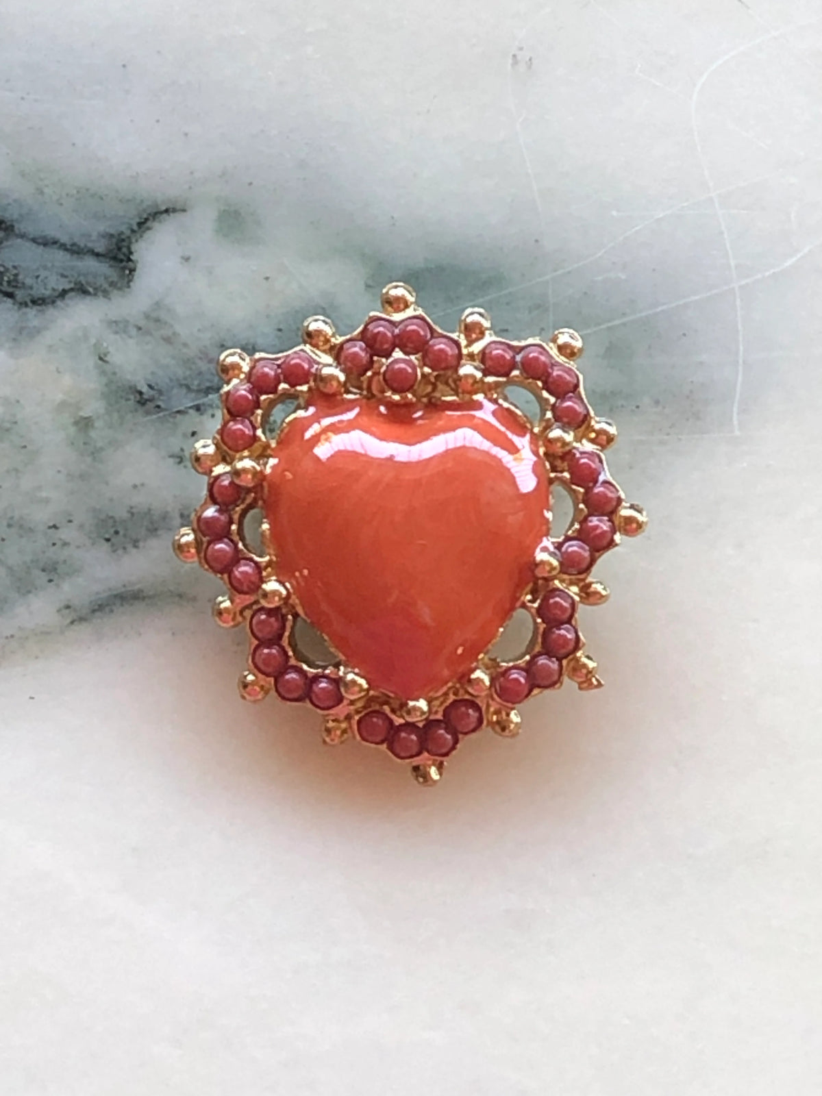 Broochella Sacro Cuore Fiorentina Heart Brooch in coral with a glossy heart centre, ornate gold-tone detailing and coral bead embellishments, photographed on a white marble background.