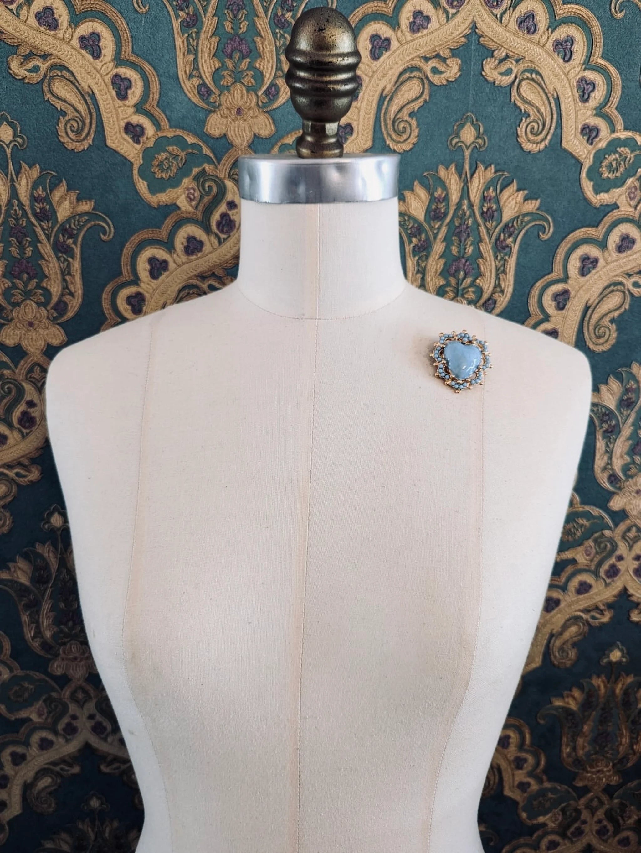 Broochella Sacro cuore fiorentina blue with gold metal base heart shaped brooch. Pinned to a mannequin. The background is green and gold damask wallpaper