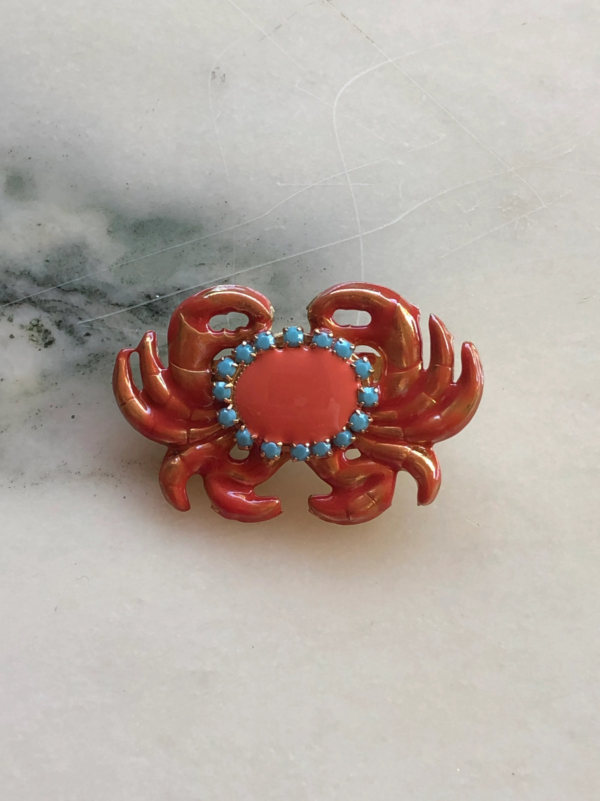 Broochella Men's Sacro Cuore Granchio Crab Brooch in glossy coral enamel with turquoise stones and gold detailing, photographed on a white marble background.