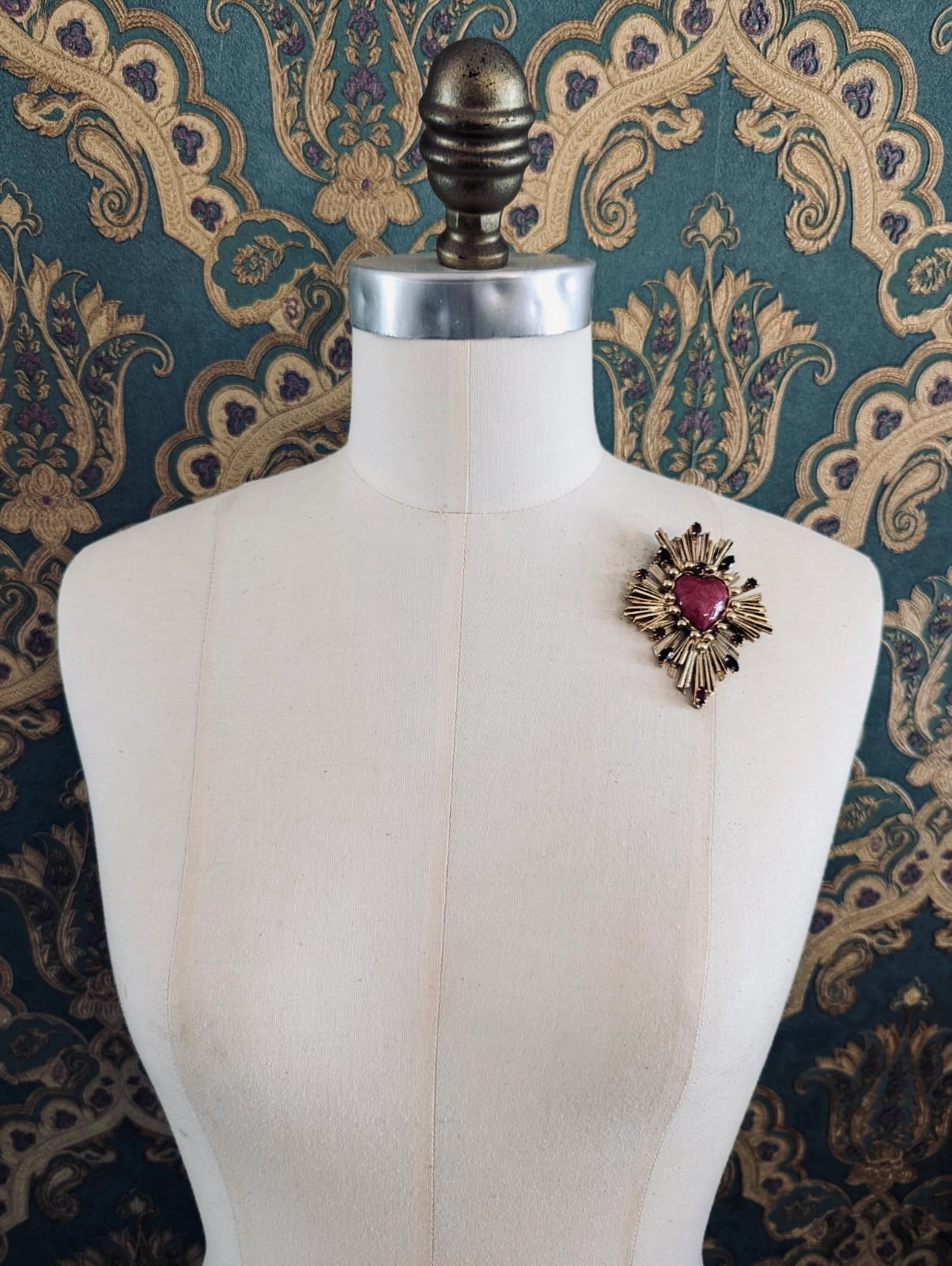 Broochella Sacro cuore lucia gold metal and red enamel heart shaped brooch. Pinned to a mannequin. The background is green and gold damask wallpaper
