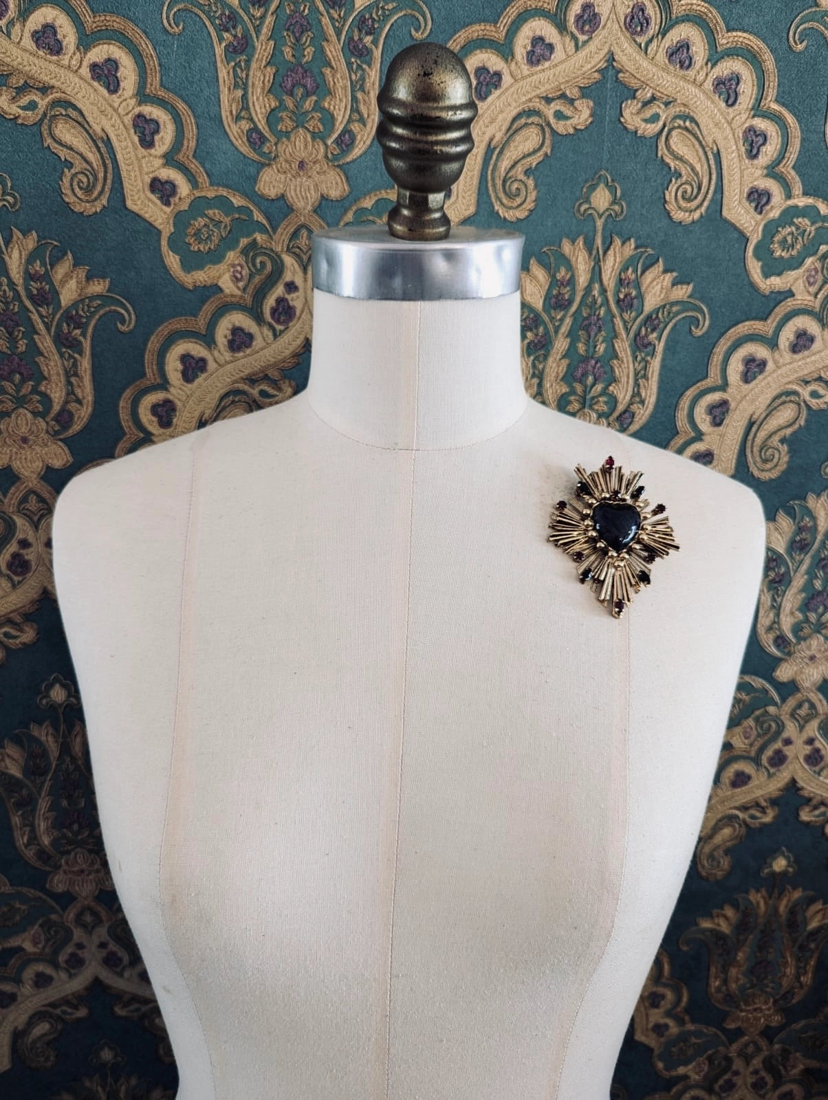 Broochella Sacro cuore lucia gold metal and red enamel heart shaped brooch. Pinned to a mannequin. The background is green and gold damask wallpaper