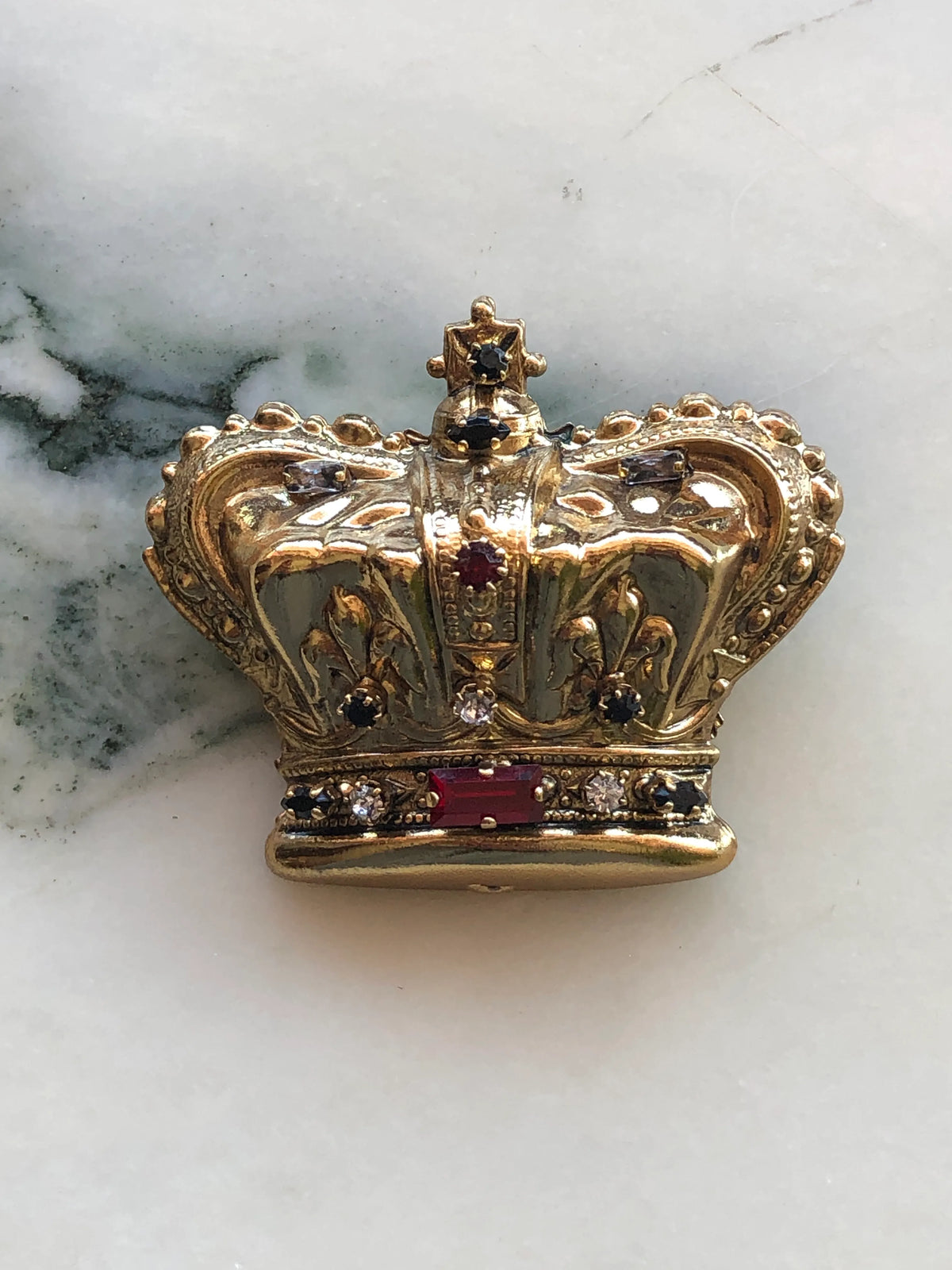 Broochella Sacro Cuore Regina Crown Brooch in antique gold with ornate detailing and red, black and clear crystal accents, photographed on a white marble background.