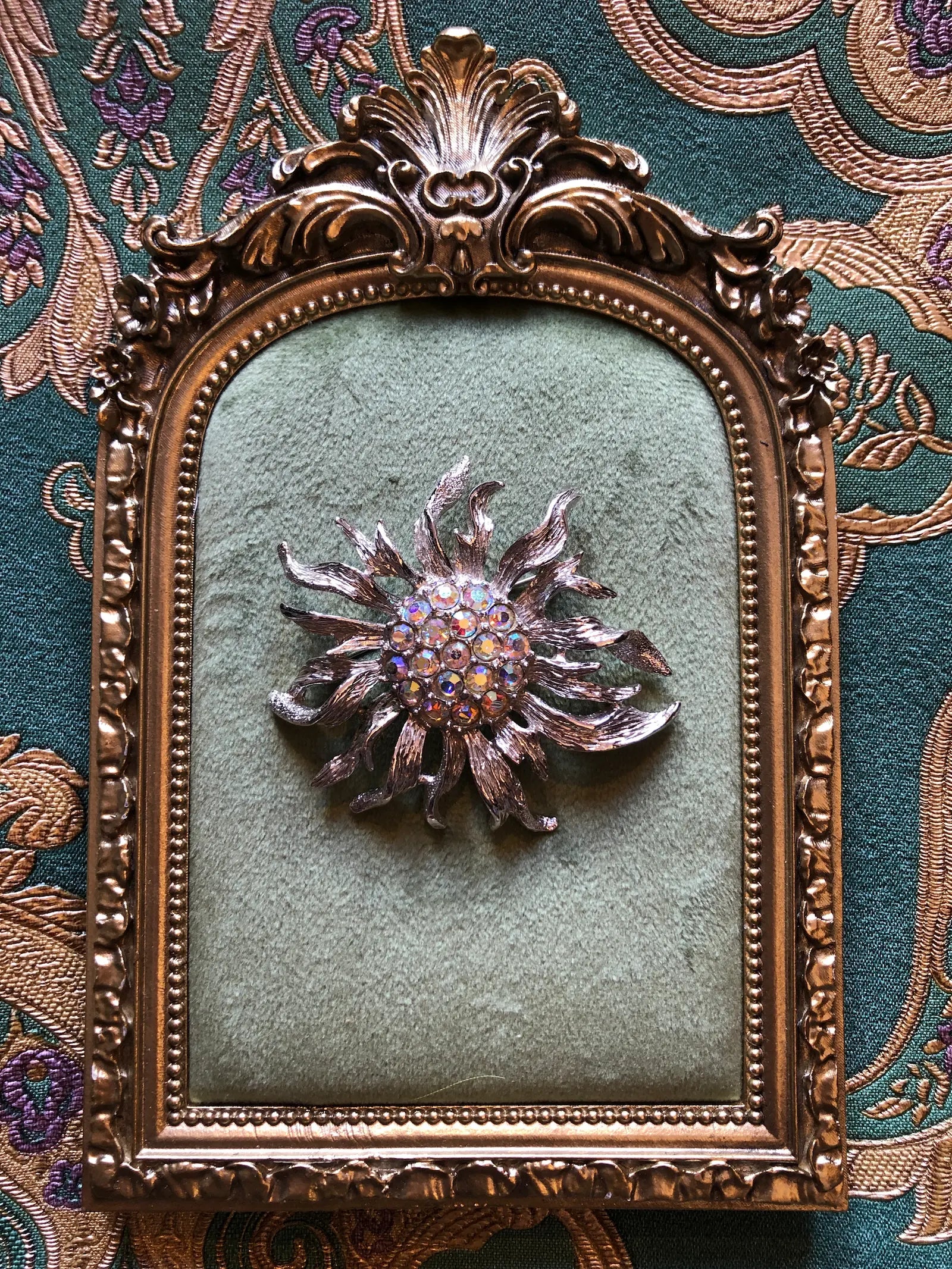A vintage Sarah Coventry brooch featuring a metallic sunburst design with a purple and gold centre, mounted on a square, textured sage green panel with an ornate gold frame, set against a patterned green and gold fabric background.