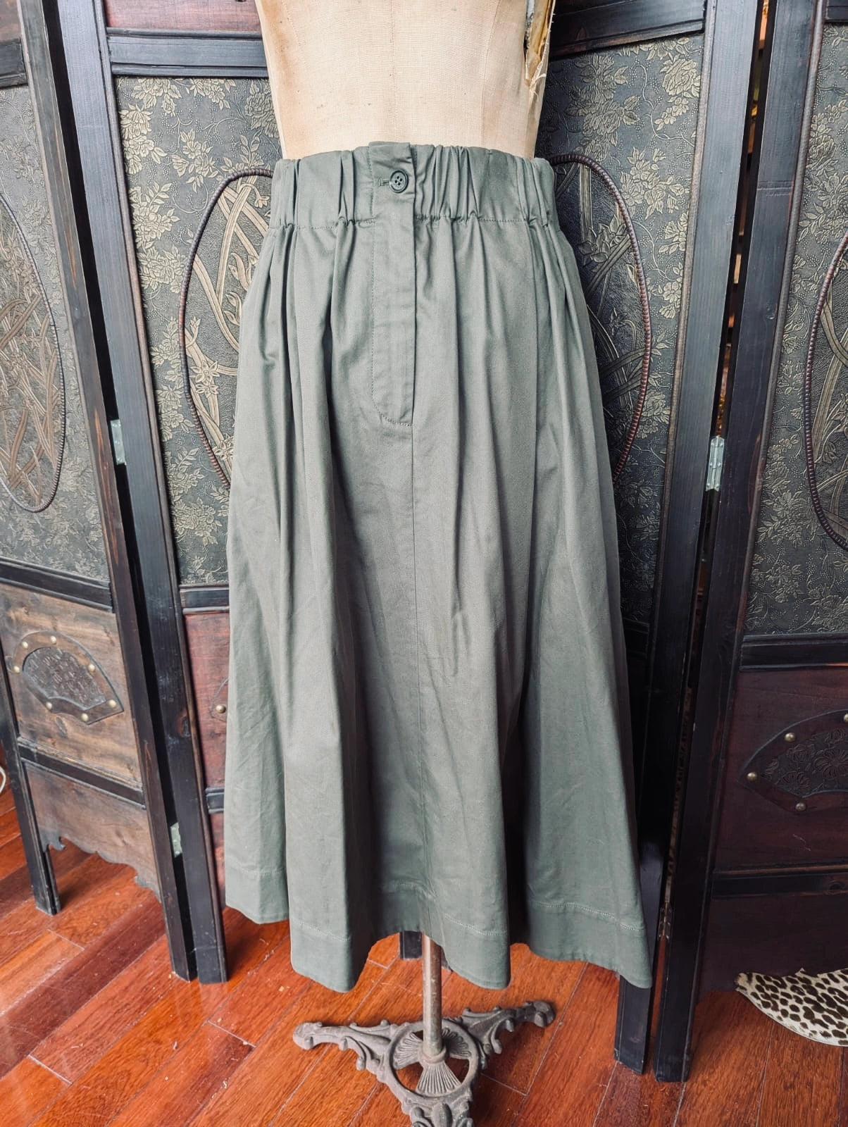 Broochella The Stylist's Closet Scanlan Theodore designer olive green high-waisted A-line skirt displayed on a dress form mannequin, set against a dark wood folding screen and wooden floor.