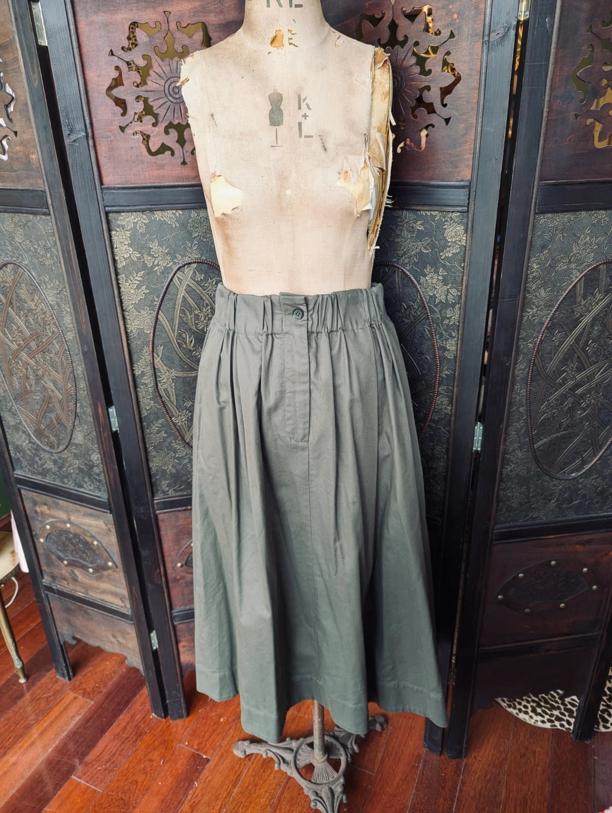 Broochella The Stylist's Closet Scanlan Theodore designer olive green high-waisted A-line skirt displayed on a dress form mannequin, set against a dark wood folding screen and wooden floor.
