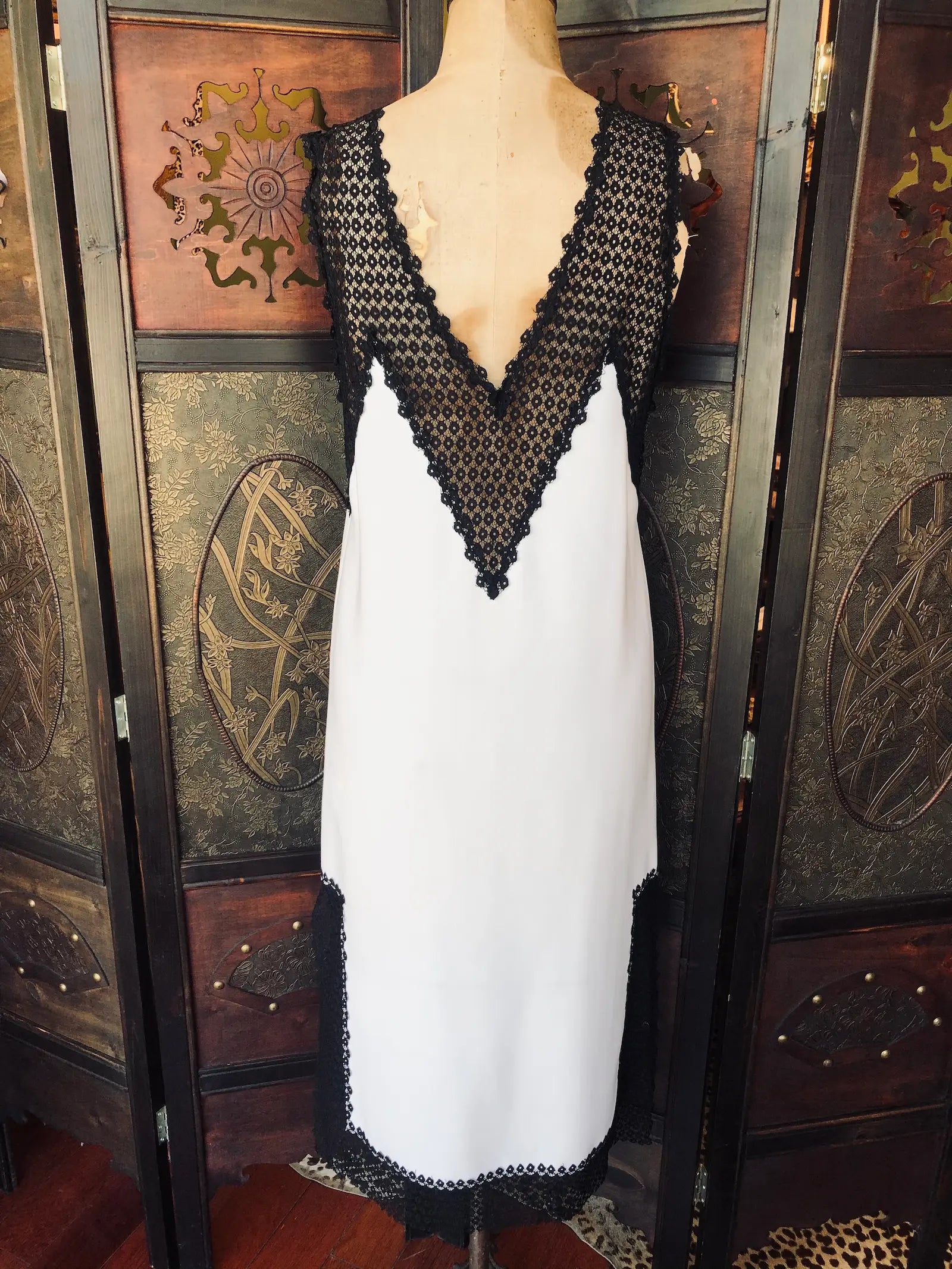 Broochella Scanlan Theodore midi slip dress. Size AU 6 It is Ivory with black lace detailing. It has Deep V lace neckline, 
sleeveless design, black lace-trimmed hem and a slip-style silhouette. It is pictured on a vintage mannequin. The background is a an ornate, dark wood, vintage room divider. 