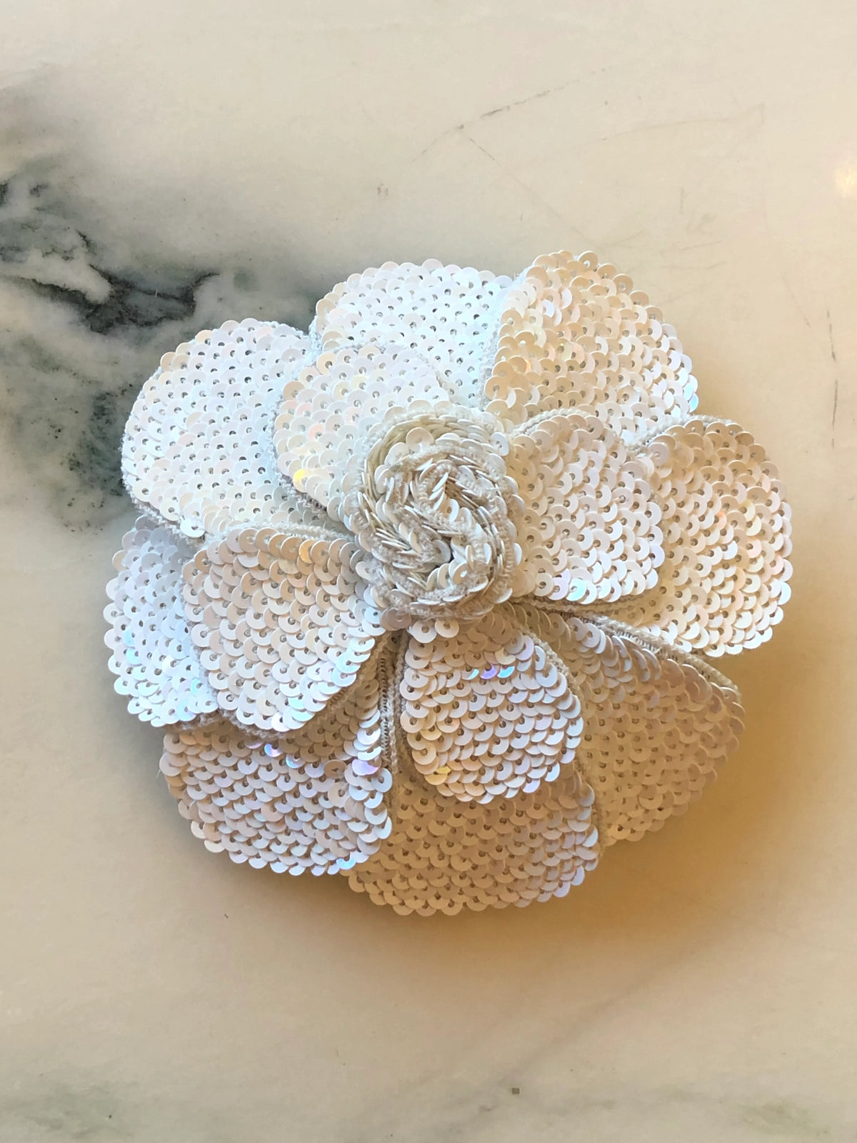 Broochella Scintillante Bridal Sequin Flower Brooch in white, featuring layered sculptural petals covered in shimmering white sequins with an intricate floral centre, photographed on a cream marble background.