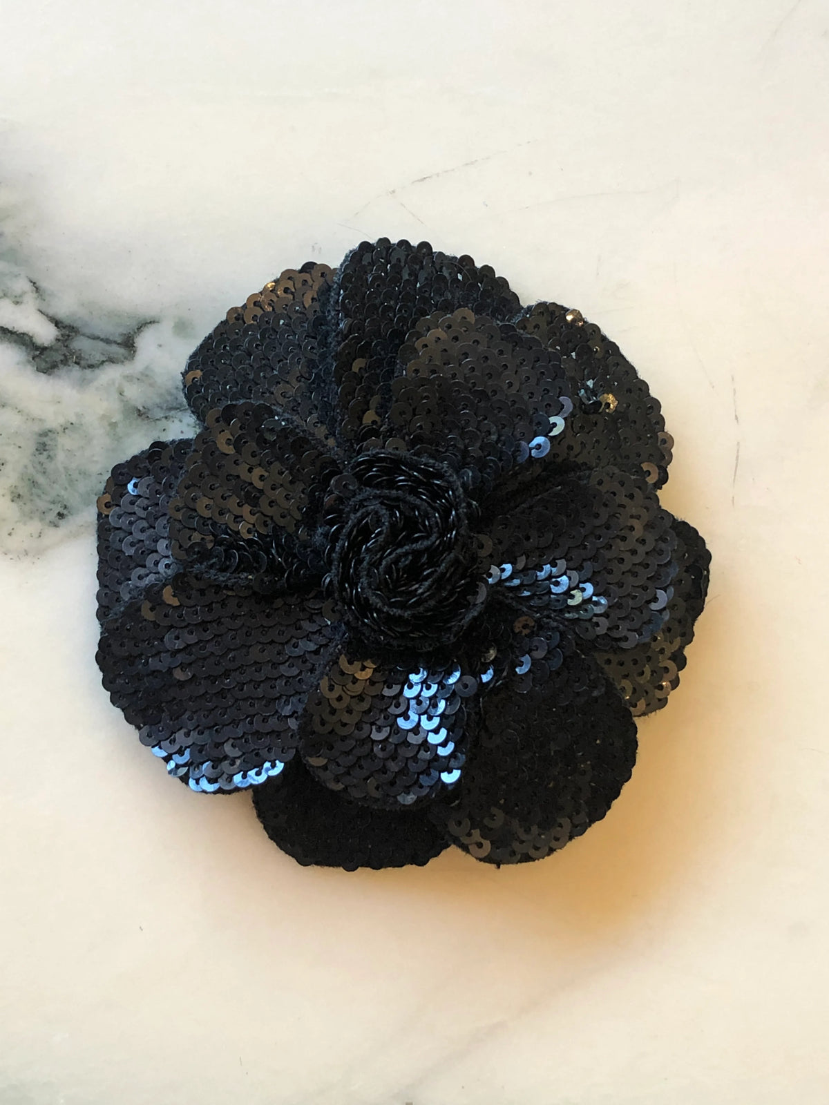Broochella Scintillante Men's Flower Brooch Pin in black, featuring layered sculptural petals embellished with tonal black sequins and a textured black centre on a cream marble background.