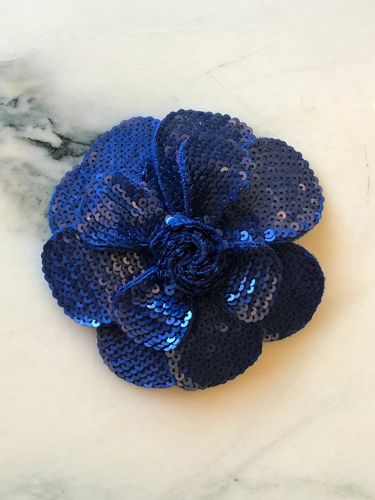 Broochella Scintillante Flower Brooch Pin in deep blue, featuring layered sculptural petals embellished with tonal navy and cobalt blue sequins and a textured blue centre on a cream marble background.