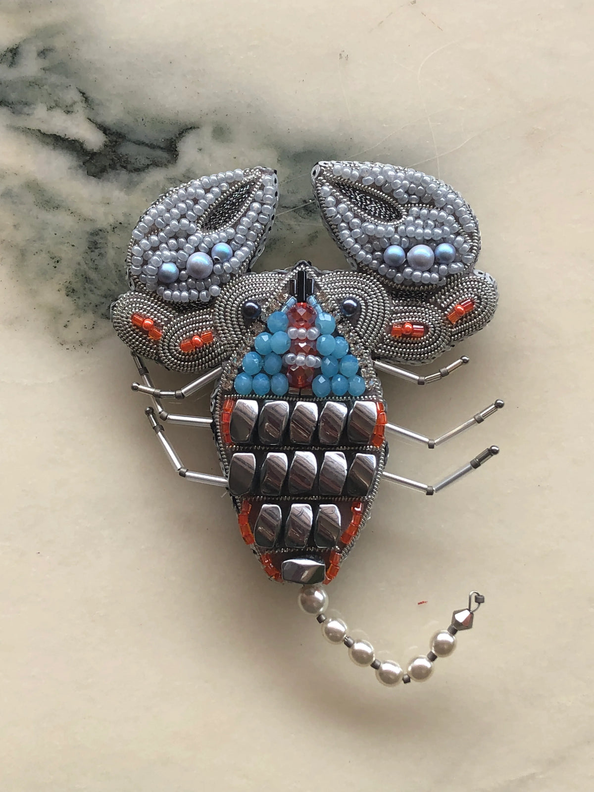 A Broochella Zodiac Scorpio scorpion-shaped beaded brooch featuring silver-toned metal mesh, blue and turquoise beads, orange-red accents, and a curved tail of silver beads, displayed on a light beige marble surface.