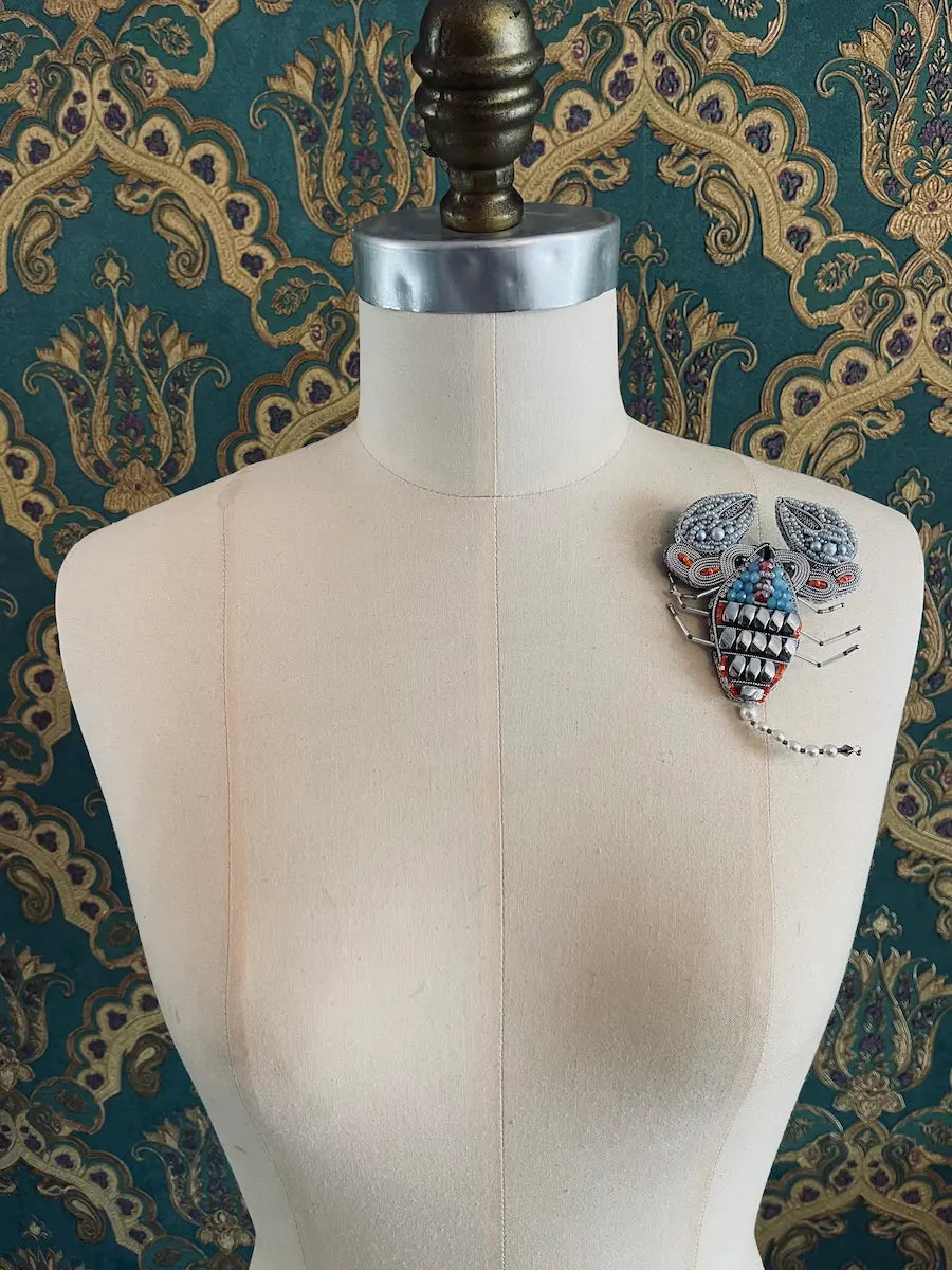 Broochella scorpio hand beaded orange silver designer zodiac brooch. Styled on a mannequin. The background is green and gold damask wallpaper