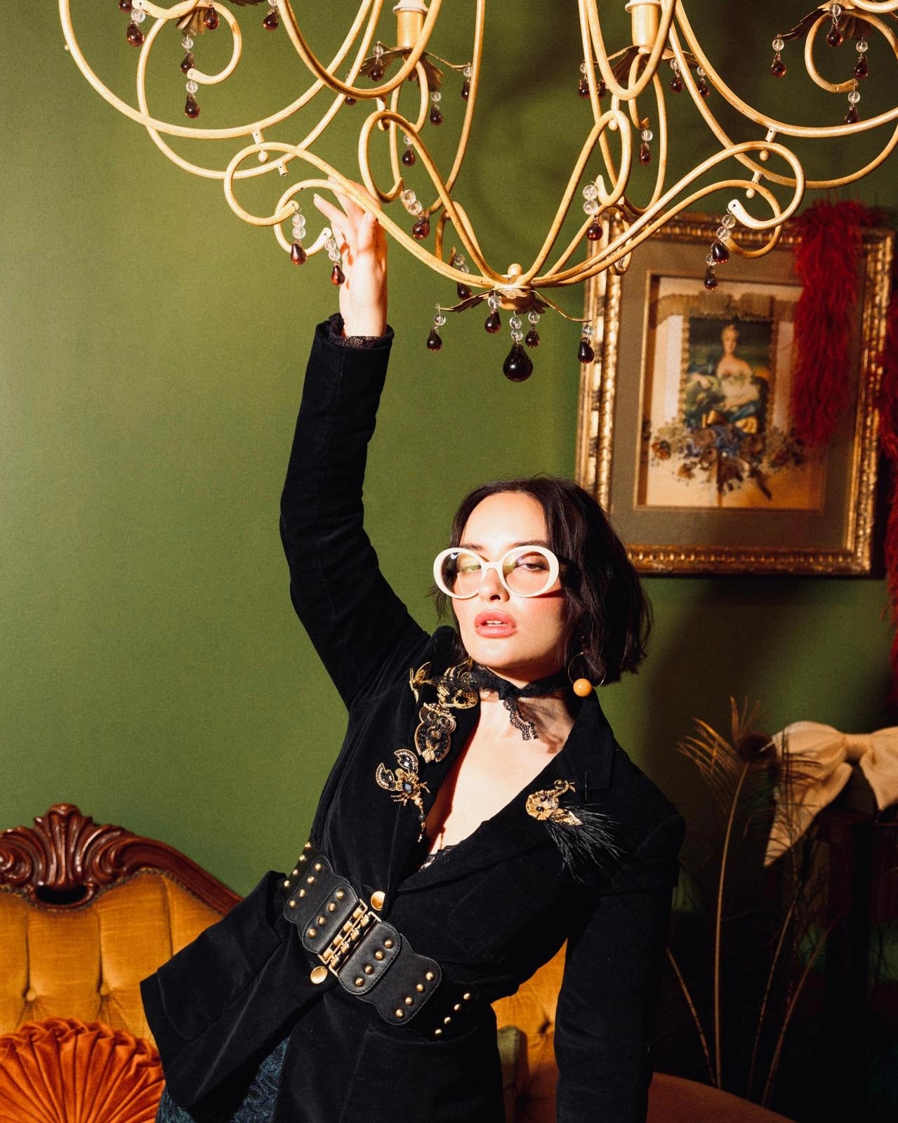 A woman wearing a black velvet jacket with ornate gold embroidery, a wide black belt with gold studs, and round white-framed glasses reaches up to touch a large gold chandelier in a vintage-style room with a green wall. She is wearing the Broochella scorpion brooch in black and gold