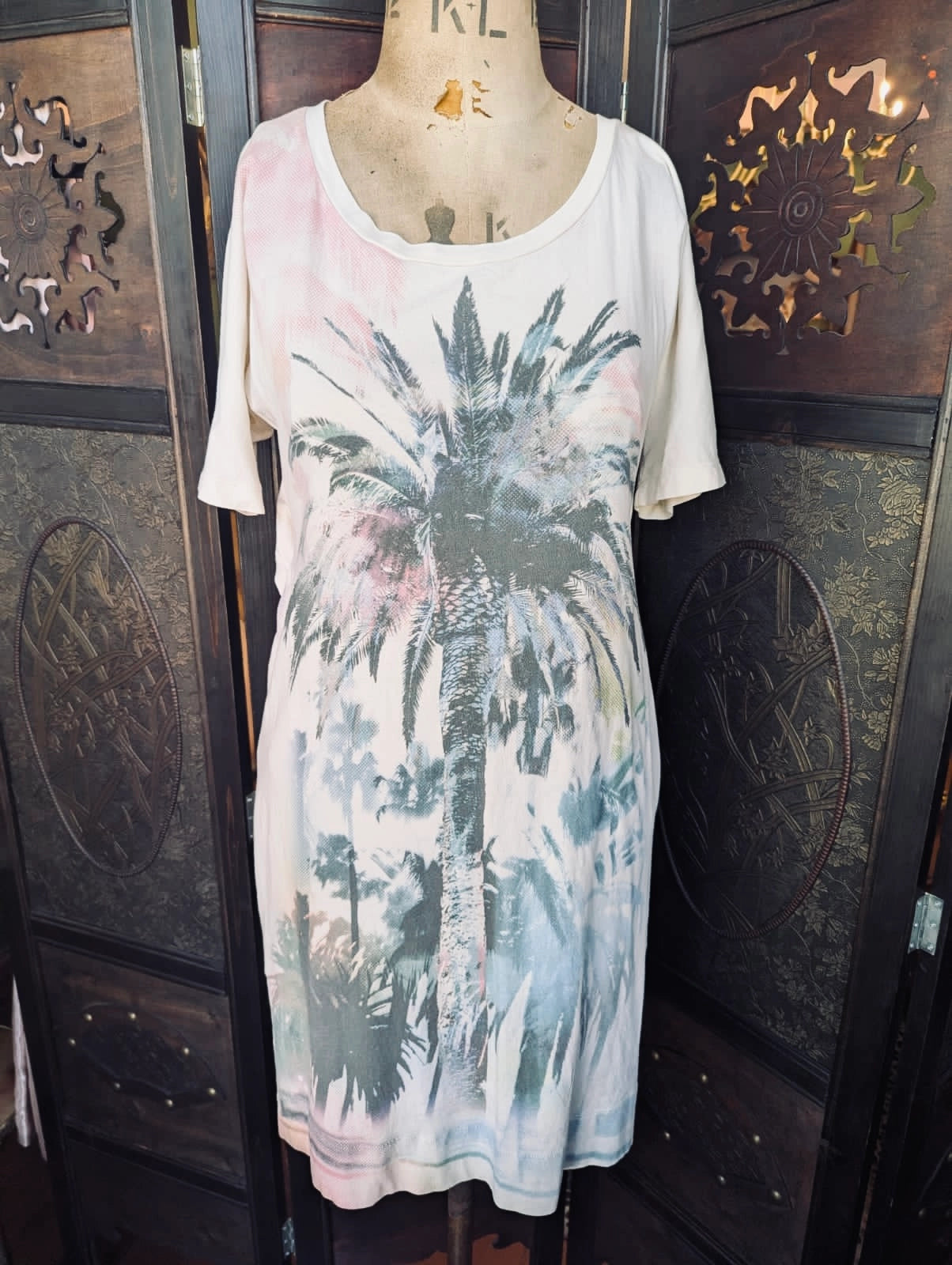 A Broochella See By Chloe short-sleeve scoop-neck t-shirt dress displayed on a mannequin torso, featuring a large palm tree graphic in shades of grey, blue, and green over a light pink and off-white base, set against a dark carved wooden screen.