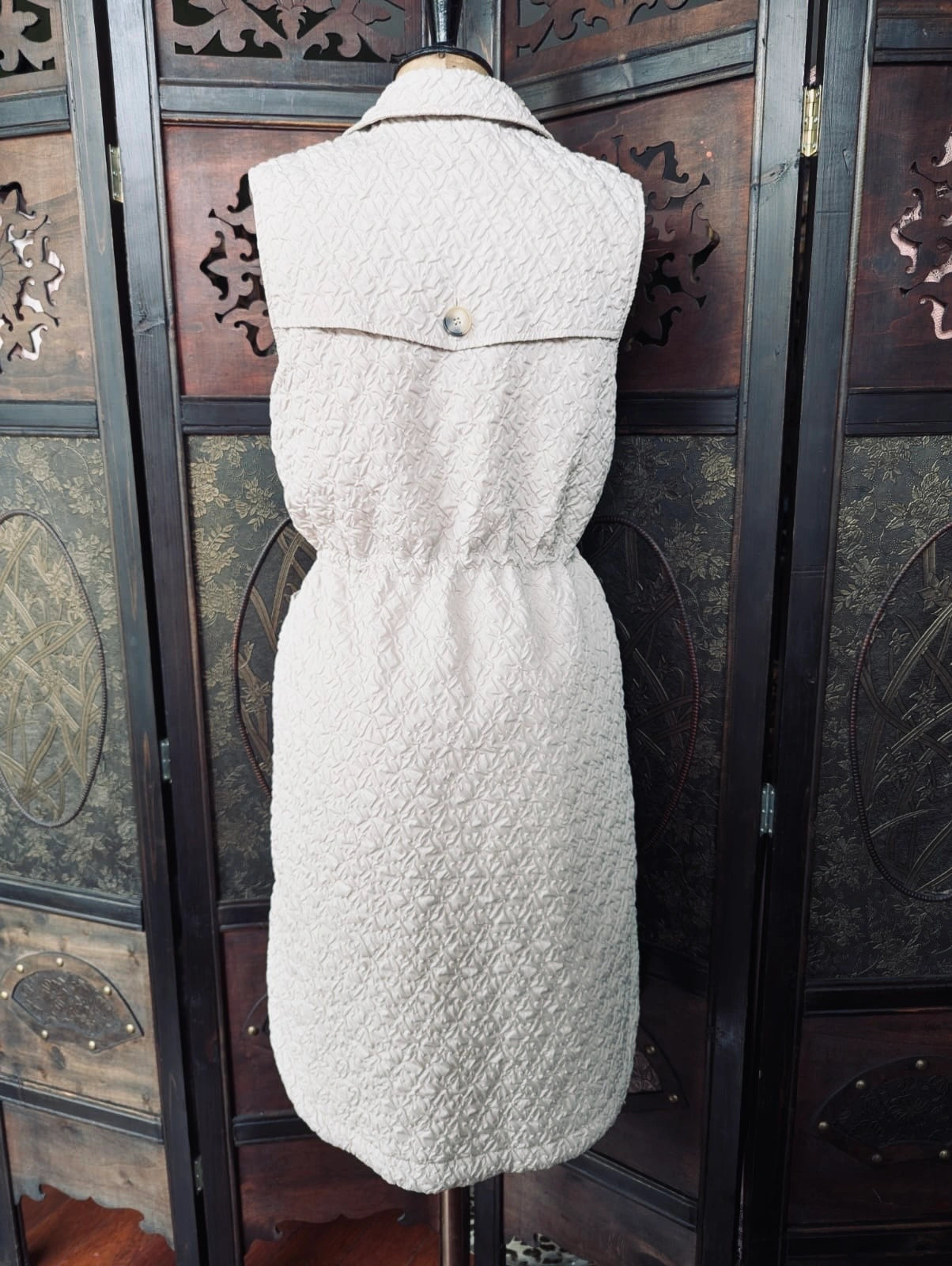 Broochella The Stylist's Closet Selected Femme longline beige quilted vest viewed from the back. It is displayed on a vintage mannequin. The background is an ornate, vintage, dark wood room divider. 