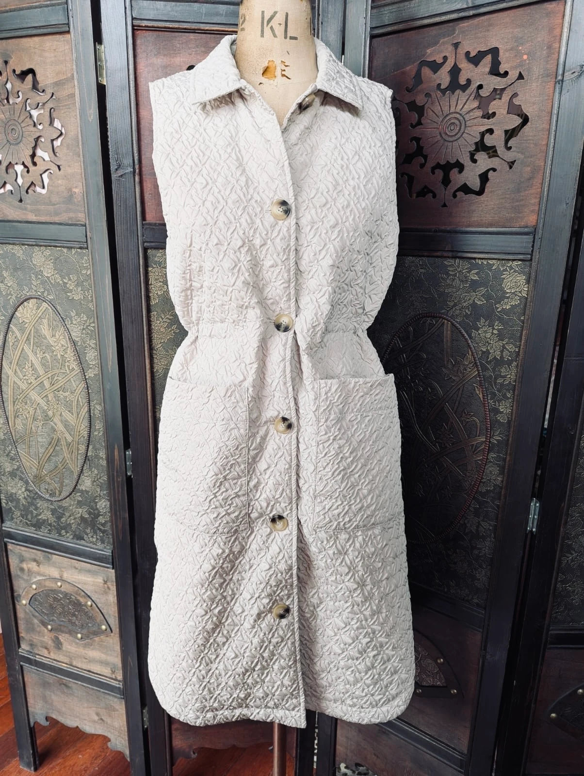 Broochella The Stylist's Closet Selected Femme longline beige quilted vest. It is displayed on a vintage mannequin. The background is an ornate, vintage, dark wood room divider. 