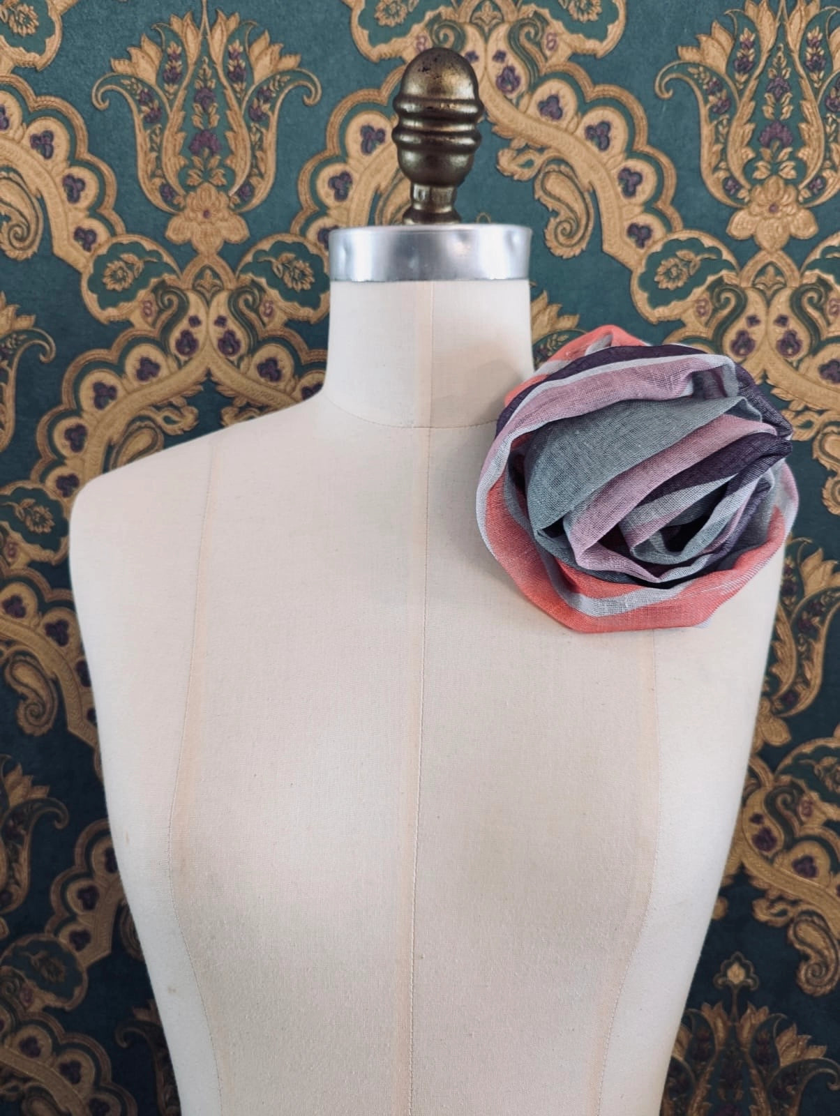 Broochella serena fiore handmade designer linen bloom brooch in grey white, pink and purple thick stripes. Pinned to a mannequin. The background is green and gold damask wallpaper.