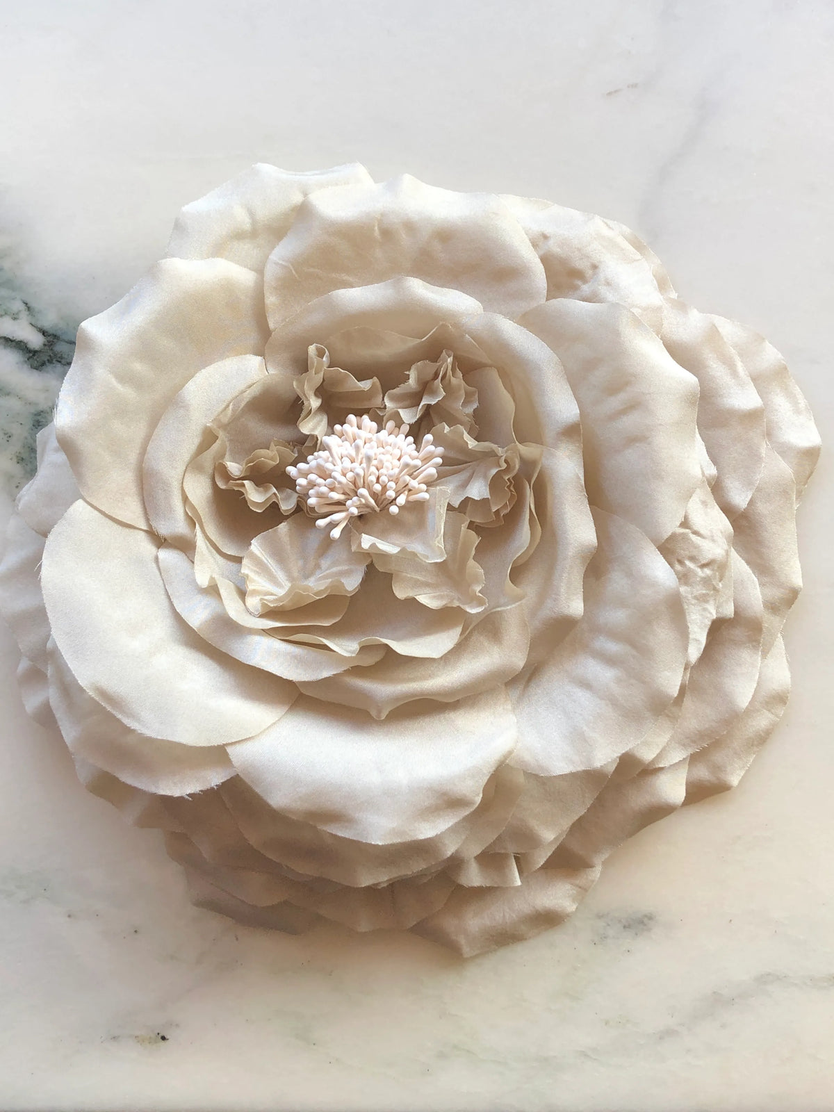 Broochella Signora Fiorita Flower Brooch Pin in soft beige with layered fabric petals and a delicate stamen centre, photographed on a white marble background with grey veining.