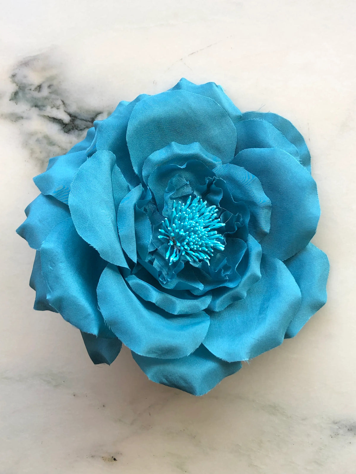 Broochella Signorina Fioritina Flower Brooch Pin in blue with layered fabric petals and a matching stamen centre, photographed on a white marble background with grey veining.