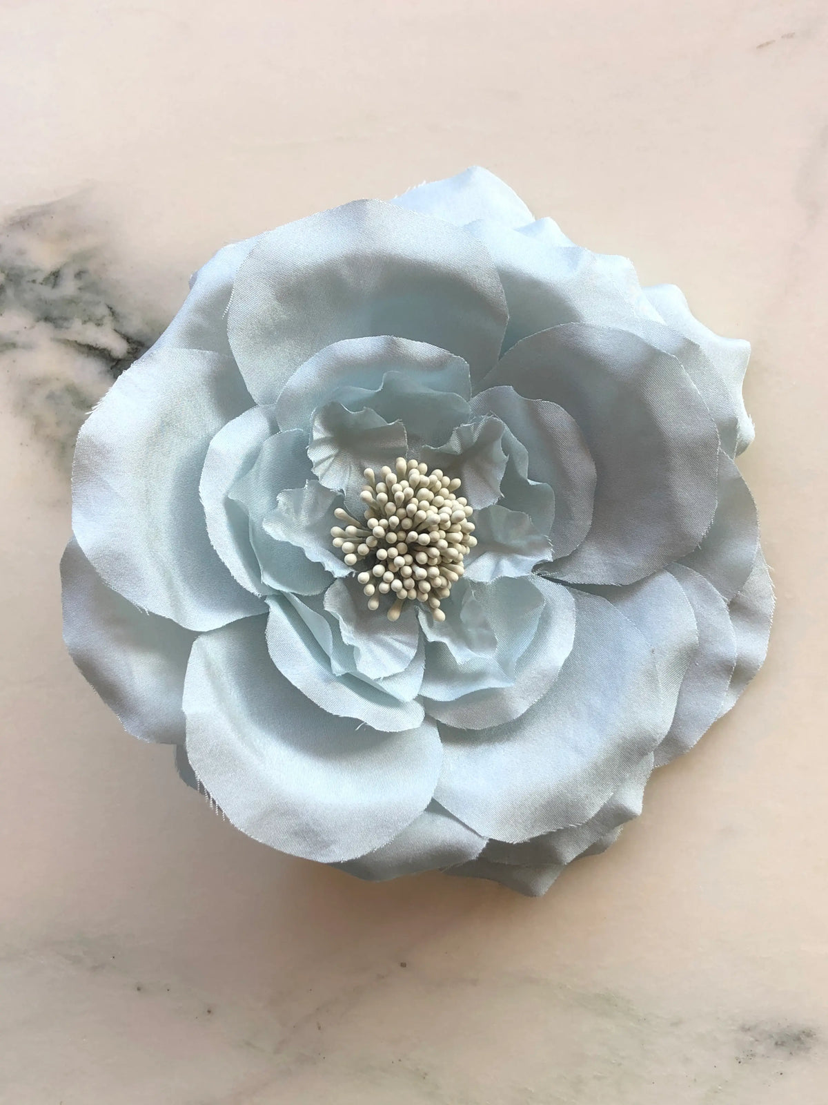 Broochella Signorina Fioritina Flower Brooch Pin in sky blue with layered fabric petals and a cream stamen centre, photographed on a white marble background with grey veining.