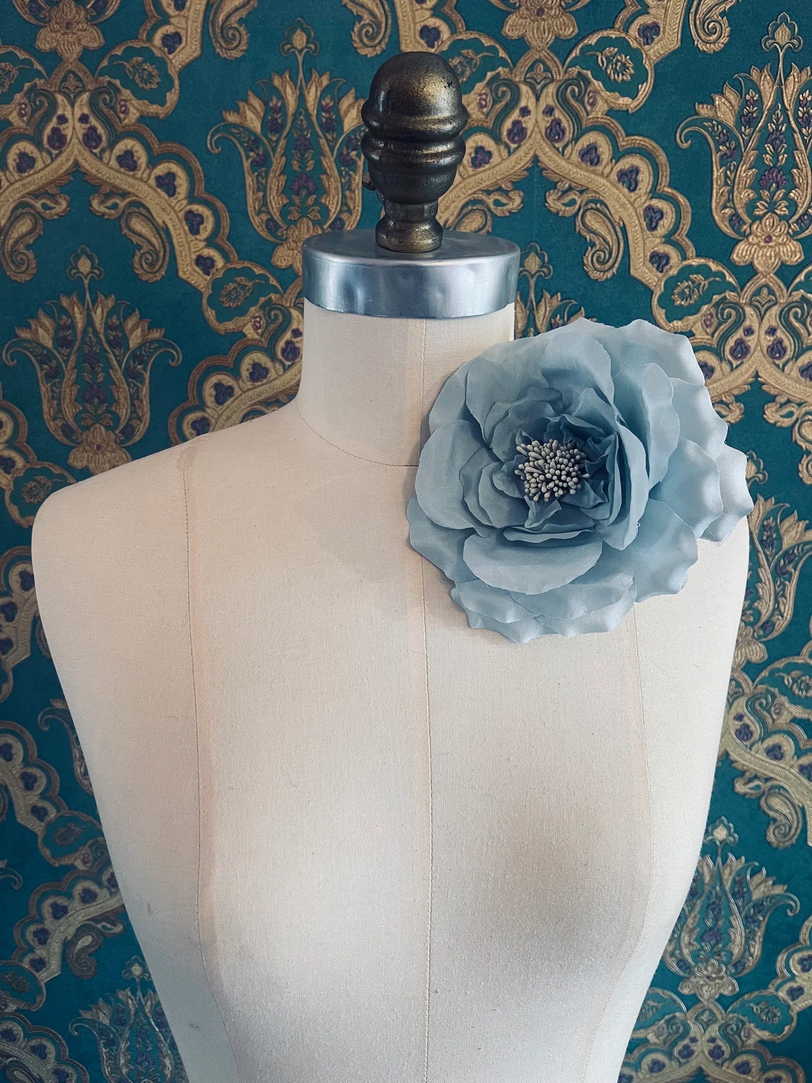 Broochella signorina fioritina handamde fabric flower brooch in sky blue. Pictured on a mannequin. The background is green and gold damask wallpaper