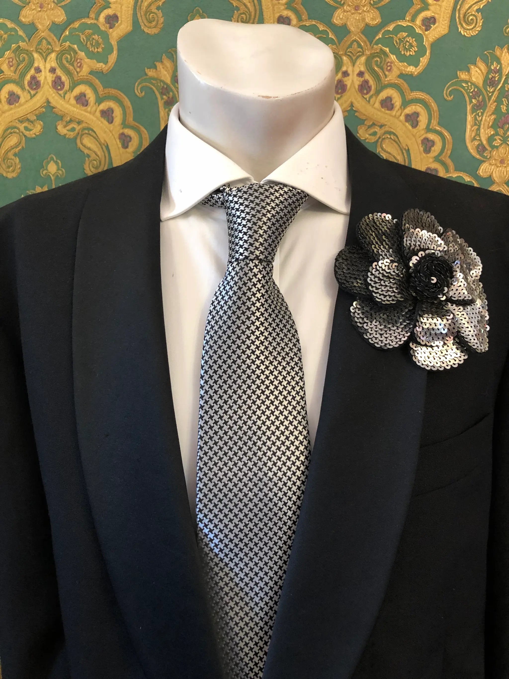A men's formal accessory set displayed on a white mannequin torso, featuring a black suit jacket, a white dress shirt, a silver and black checkered tie, and an orange Men's Broochella Scintillante sequinned flower brooch lapel pin against a green and gold patterned wallpaper background.