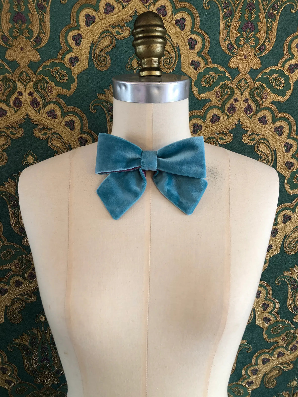 Broochella handmade Small Velvet Bow Brooch in aqua blue velvet with purple edging, styled on a cream mannequin against ornate green and gold wallpaper.