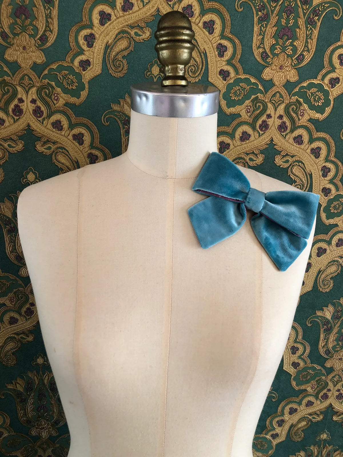 Broochella handmade Small Velvet Bow Brooch in aqua blue velvet with purple edging, styled on a cream mannequin against ornate green and gold wallpaper.