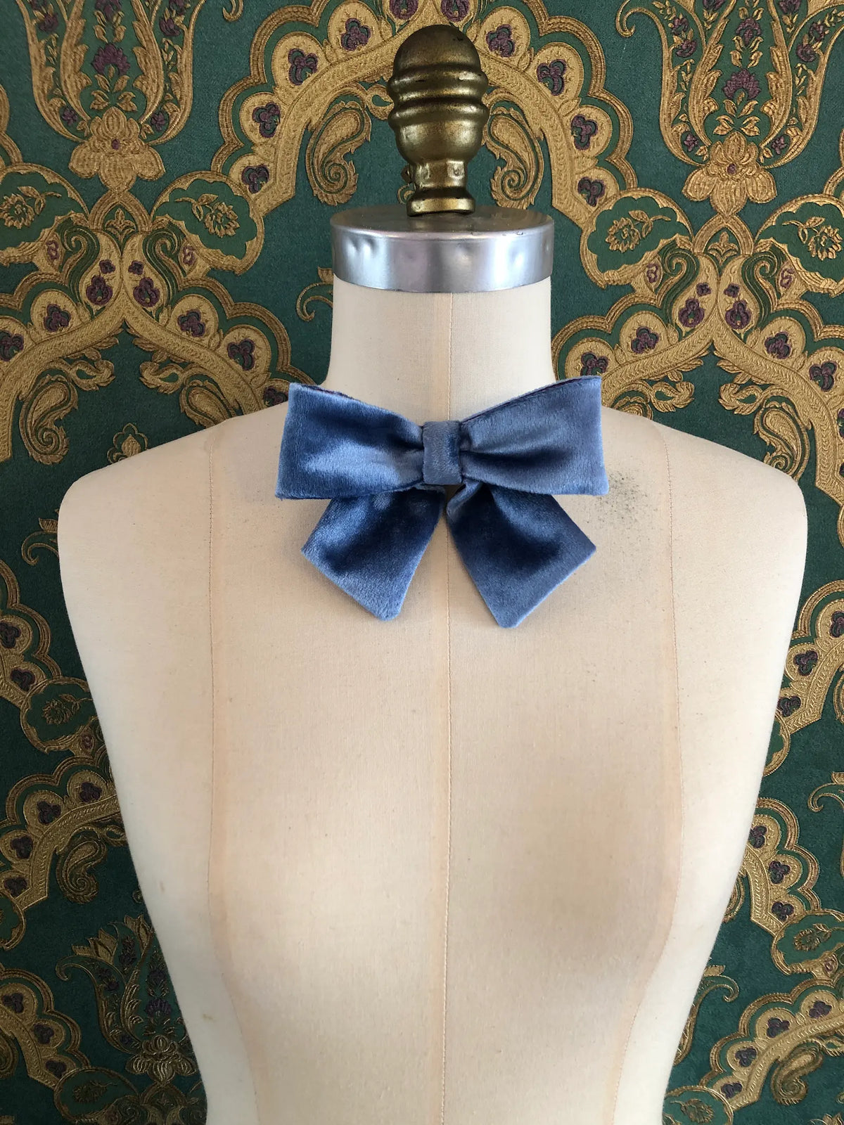 Broochella handmade Small Velvet Bow Brooch in soft blue velvet with contrasting purple edging, styled on the shoulder of a cream mannequin against ornate green and gold wallpaper.
