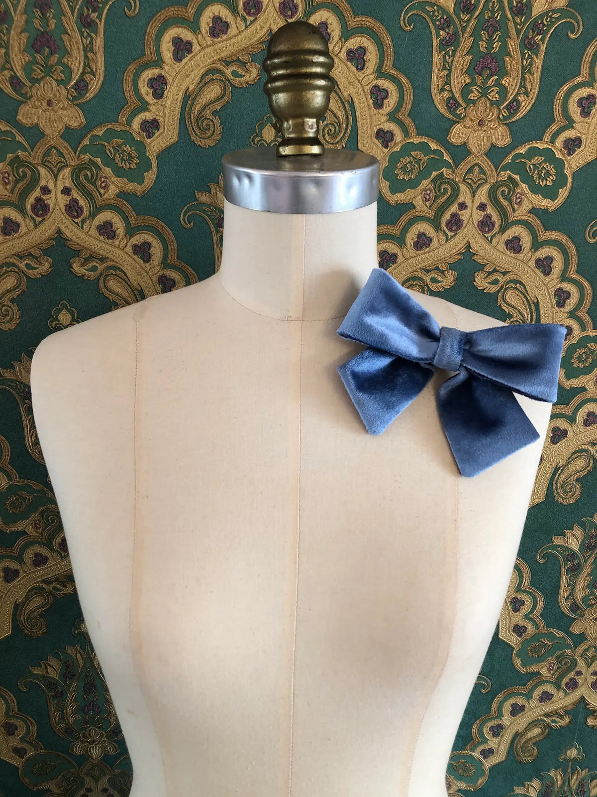 Broochella handmade Small Velvet Bow Brooch in soft blue velvet with contrasting purple edging, styled on the shoulder of a cream mannequin against ornate green and gold wallpaper.