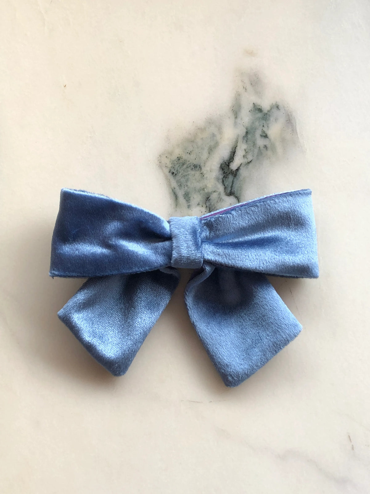 Broochella handmade men's Small Velvet Bow Brooch in soft blue velvet with contrasting purple edging, featuring a designer double bow with softly structured loops, photographed on white marble.