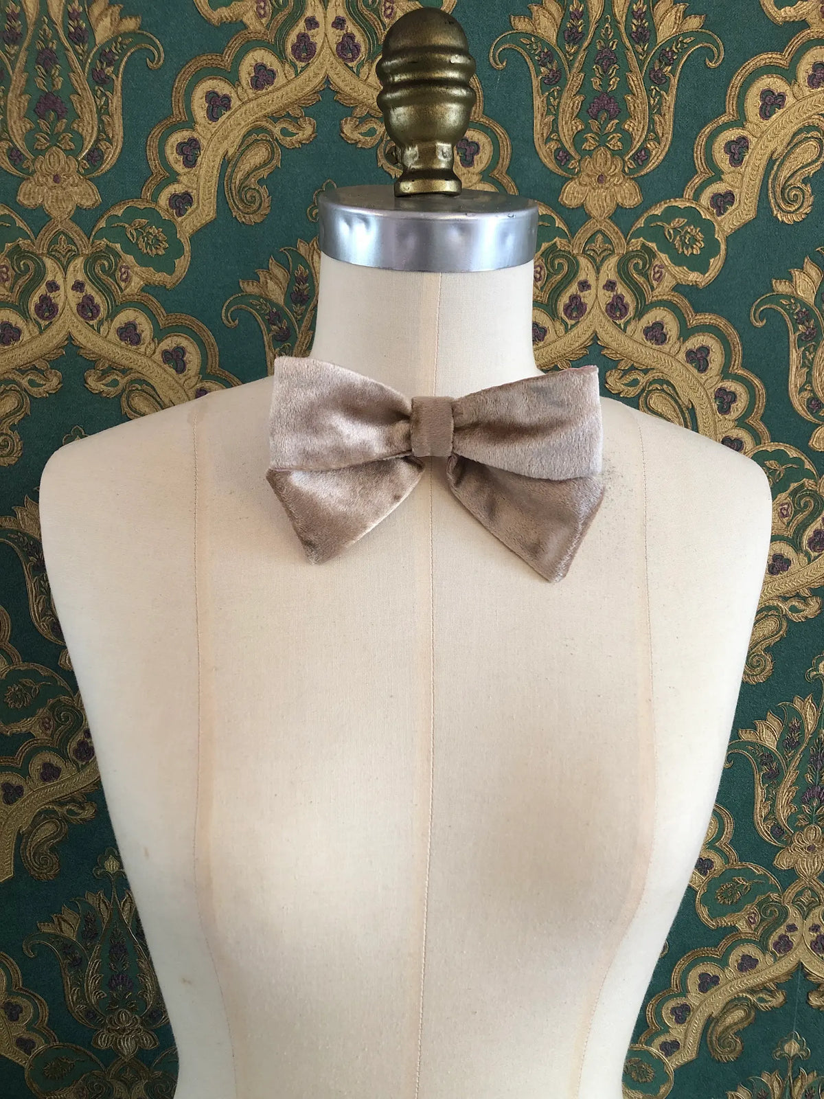 Broochella handmade Small Velvet Bow Brooch in luxurious champagne velvet with contrasting pink lining, styled on the shoulder of a cream mannequin against ornate green and gold wallpaper.