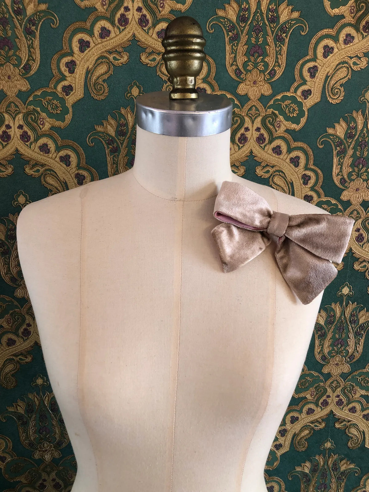 Broochella handmade Small Velvet Bow Brooch in luxurious champagne velvet with contrasting pink lining, styled on the shoulder of a cream mannequin against ornate green and gold wallpaper.