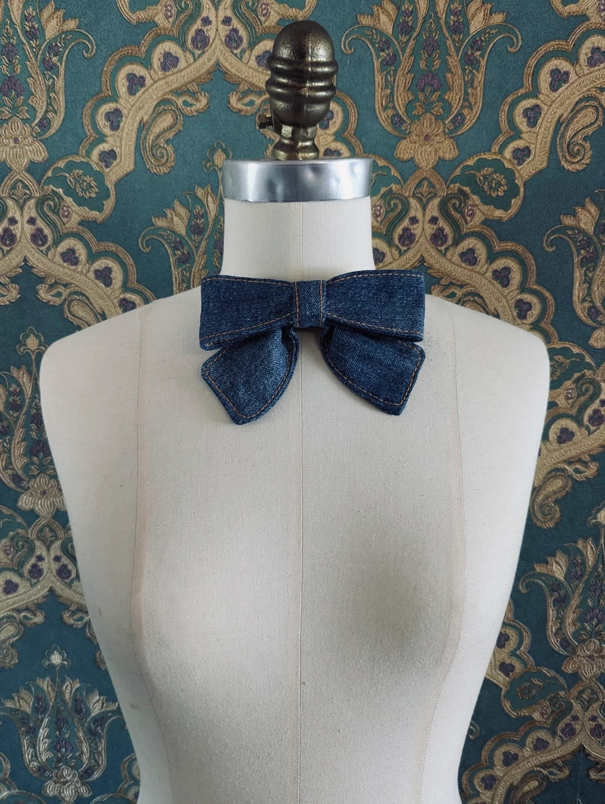 Broochella handmade blue small denim bow brooch. Up-cycled form vintage denim jeans. It is pinned to a mannequin. The background is green and gold damask wallpaper. 