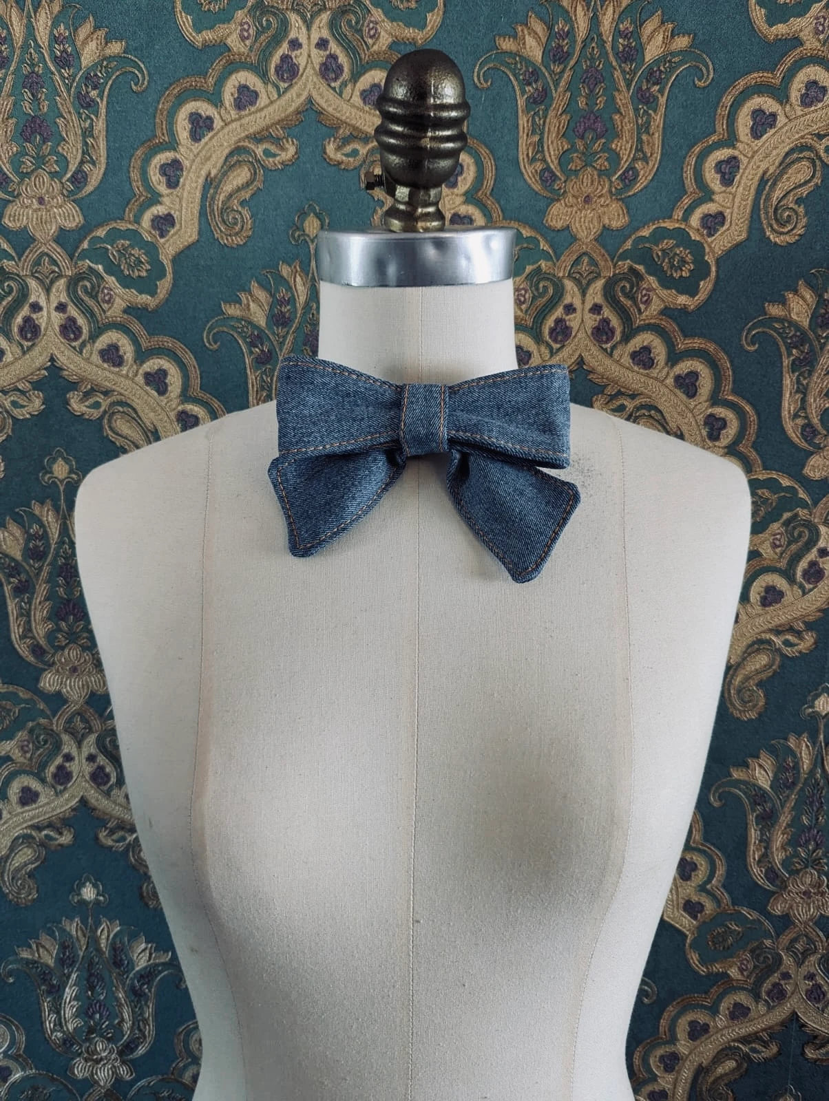 Broochella handmade sky blue small denim bow brooch. Up-cycled form vintage denim jeans. It is pinned to a mannequin. The background is green and gold damask wallpaper. 