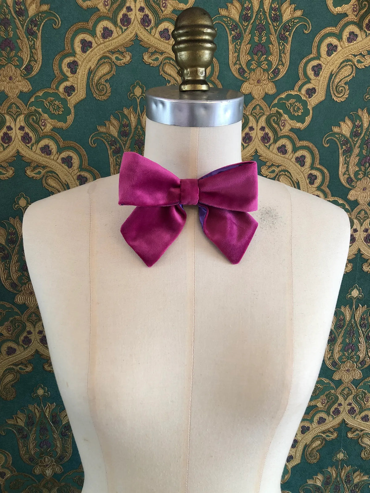 Broochella handmade Small Velvet Bow Brooch in rich magenta velvet with contrasting purple edging, styled on the shoulder of a cream mannequin against ornate green and gold wallpaper.