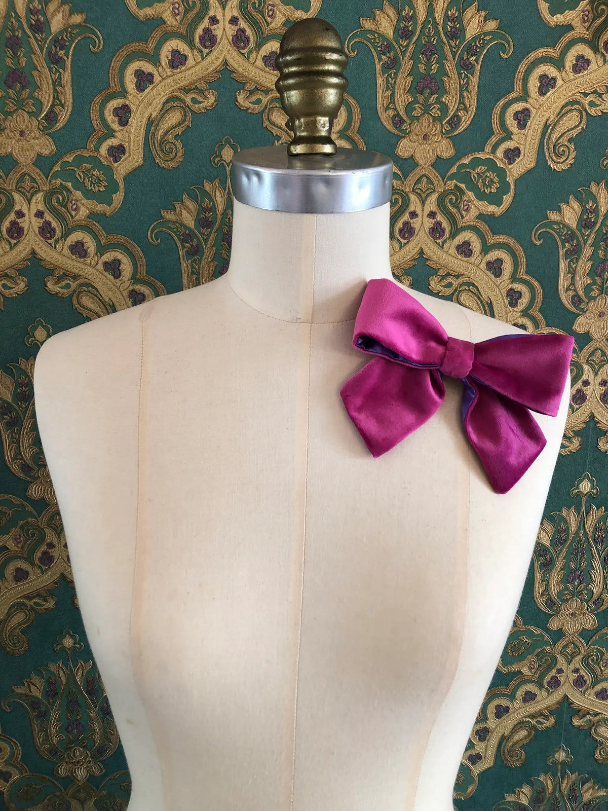Broochella handmade Small Velvet Bow Brooch in rich magenta velvet with contrasting purple edging, styled on the shoulder of a cream mannequin against ornate green and gold wallpaper.