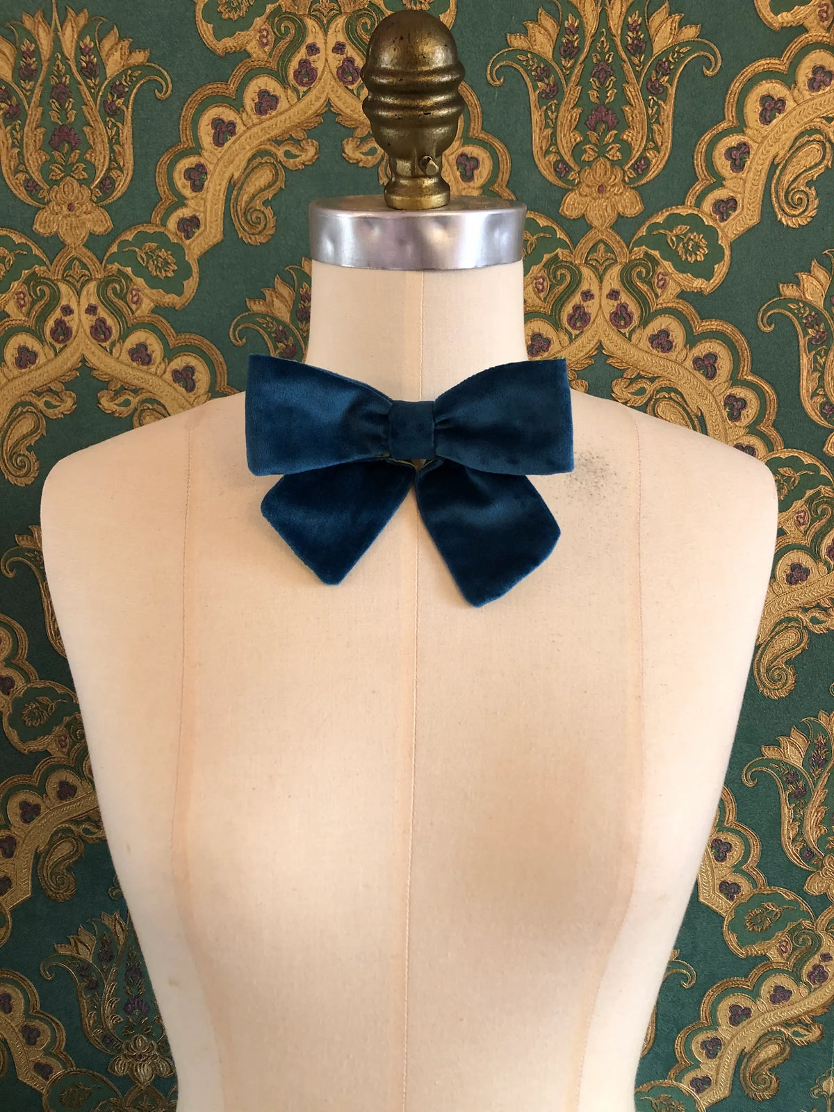 Broochella handmade Small Velvet Bow Brooch in rich peacock blue velvet with contrasting lime green lining, styled on the shoulder of a cream mannequin against ornate green and gold wallpaper.