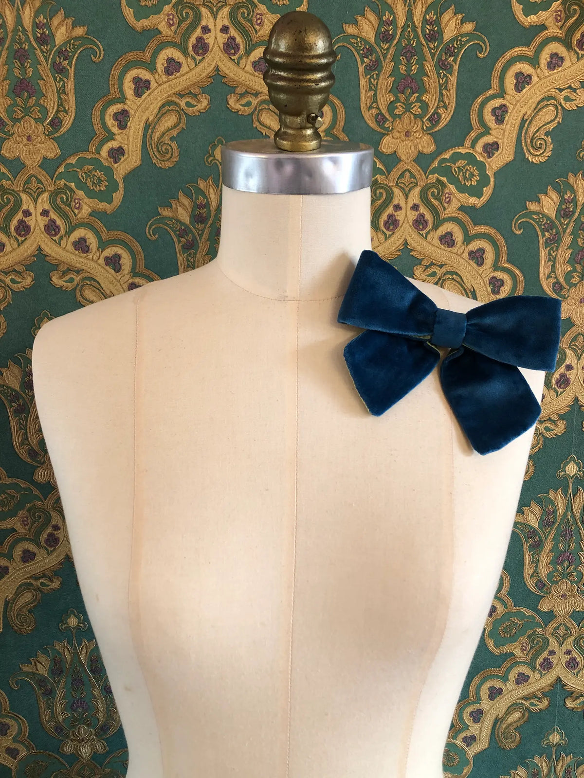 Broochella handmade Small Velvet Bow Brooch in rich peacock blue velvet with contrasting lime green lining, styled on the shoulder of a cream mannequin against ornate green and gold wallpaper.