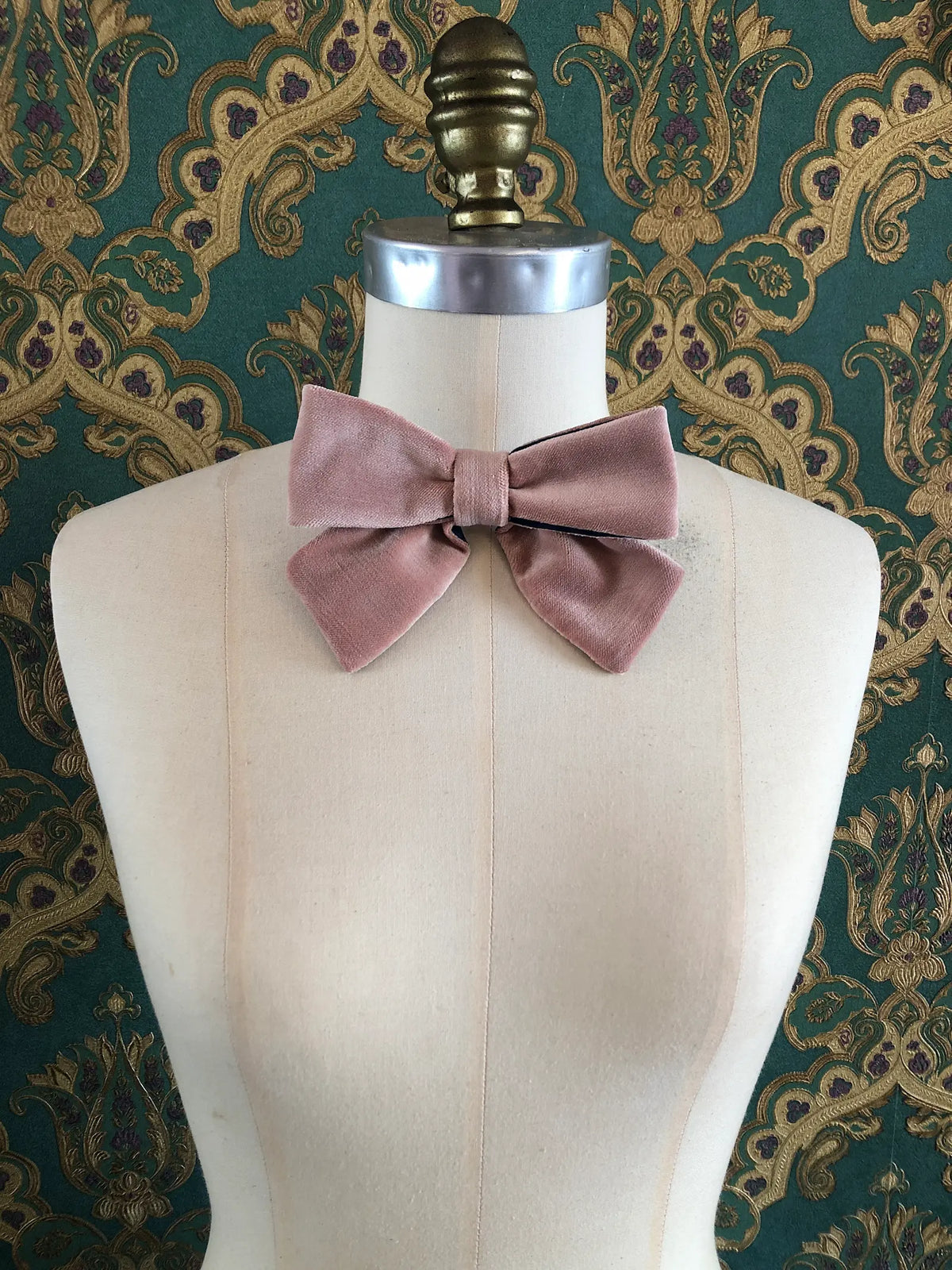 Broochella handmade Small Velvet Bow Brooch in soft blush pink velvet with contrasting navy lining, styled on the shoulder of a cream mannequin against ornate green and gold wallpaper.