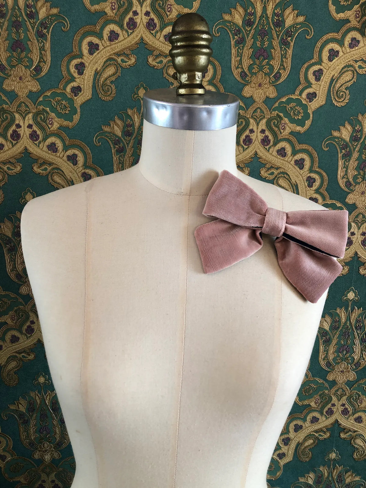 Broochella handmade Small Velvet Bow Brooch in soft blush pink velvet with contrasting navy lining, styled on the shoulder of a cream mannequin against ornate green and gold wallpaper.