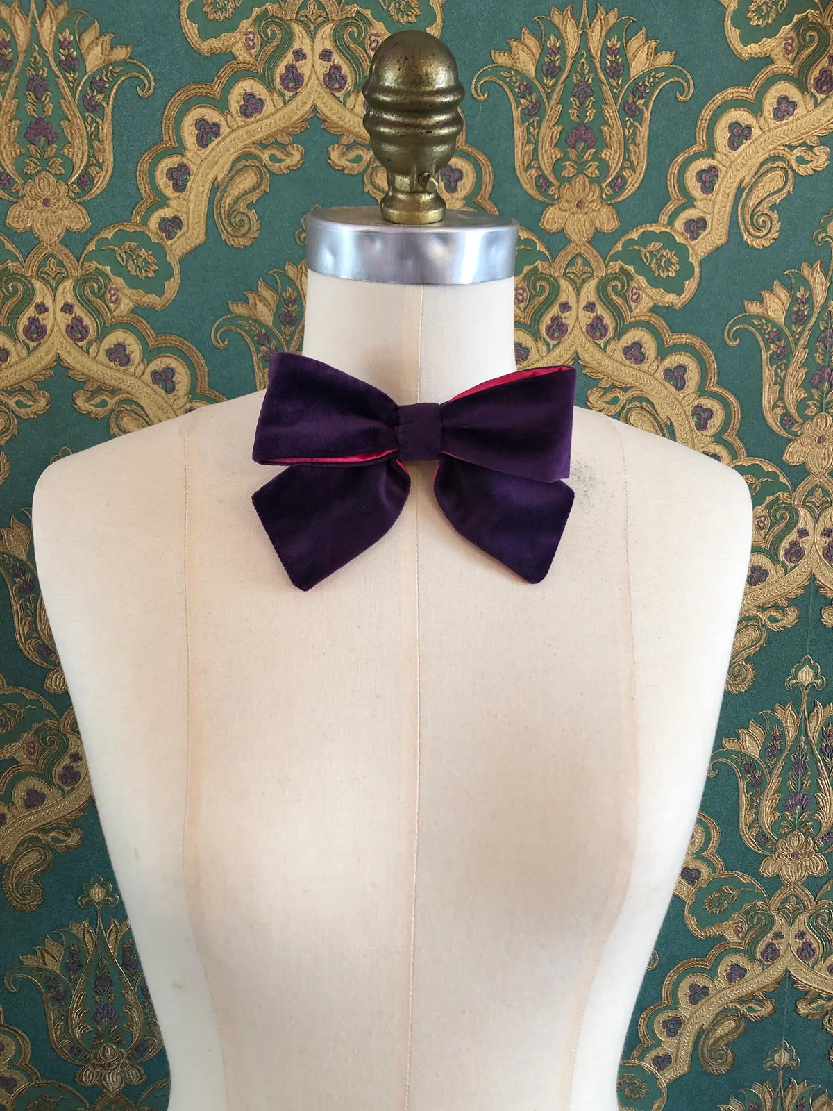Broochella handmade Small Velvet Bow Brooch in deep purple velvet with contrasting fuchsia lining, styled on the shoulder of a cream mannequin against ornate green and gold wallpaper.